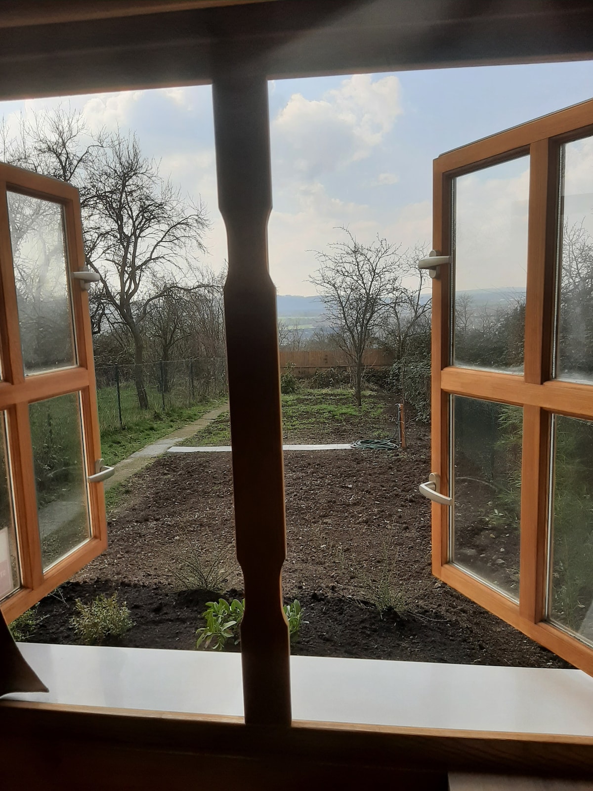Open wooden windows frame a scenic view of a garden with newly cultivated soil and sparse trees. A pathway leads through the greenery, while clouds drift in a blue sky, enhancing the tranquil atmosphere of the outdoor space.
