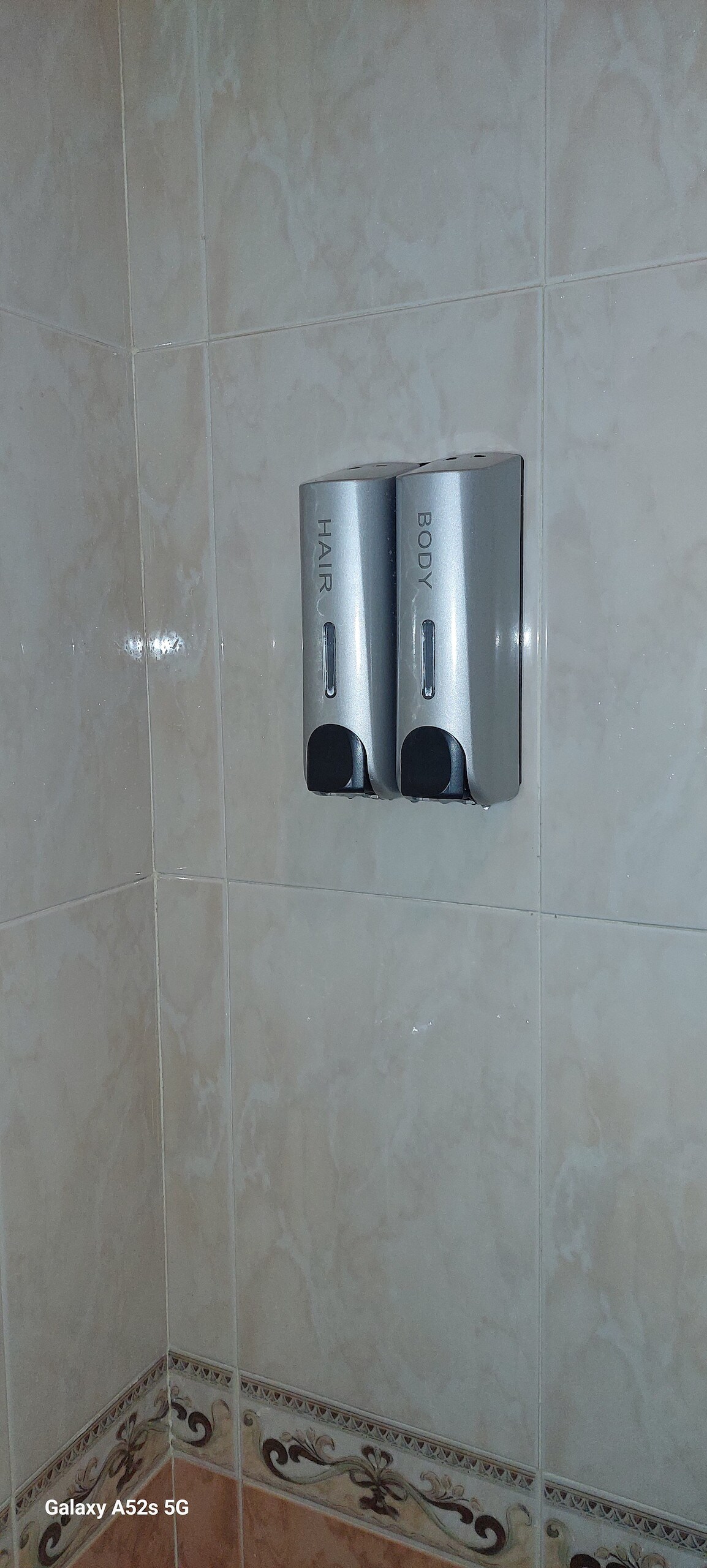A wall-mounted dispenser is visible in the shower area, featuring two compartments labelled for hair and body care products. The tiles surrounding the dispenser are light-colored with a subtle pattern, contributing to a clean and functional bathroom design.
