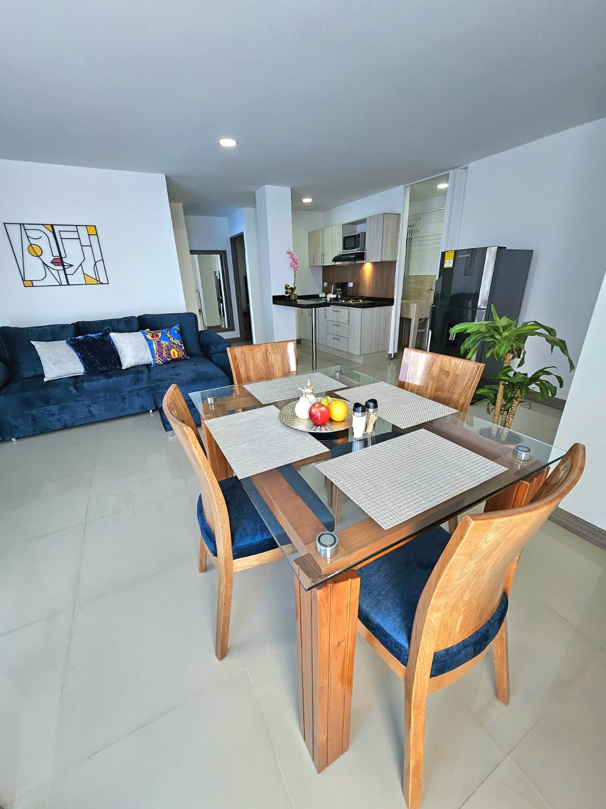 A dining area features a wooden table surrounded by four chairs, set with a decorative centerpiece. A cozy blue sofa is positioned nearby, enhancing the open layout. The background shows a glimpse of the kitchen area and a potted plant, creating a welcoming atmosphere.