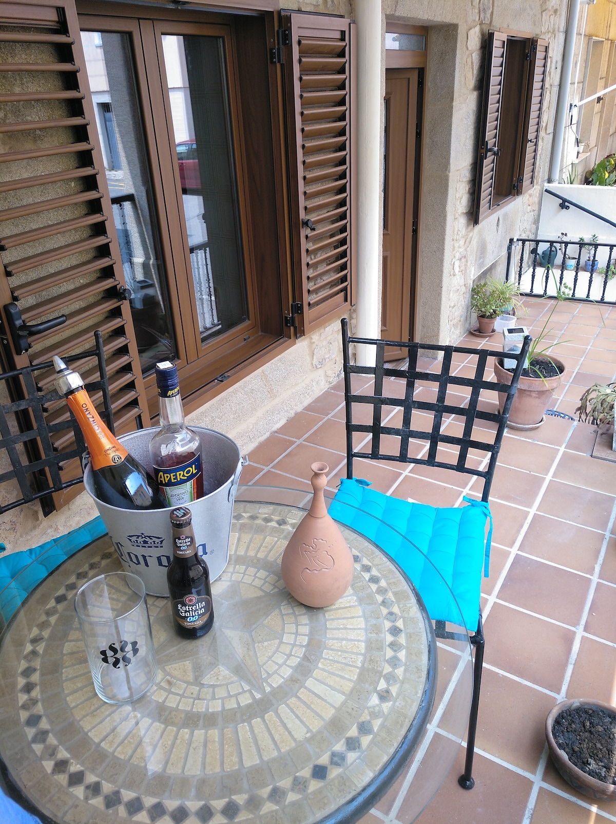 A table set outdoors features a decorative mosaic top, complemented by a metal chair. Various beverage bottles rest in an ice bucket, alongside a clay pitcher and a glass. Louvered windows provide natural light, with potted plants enriching the space.