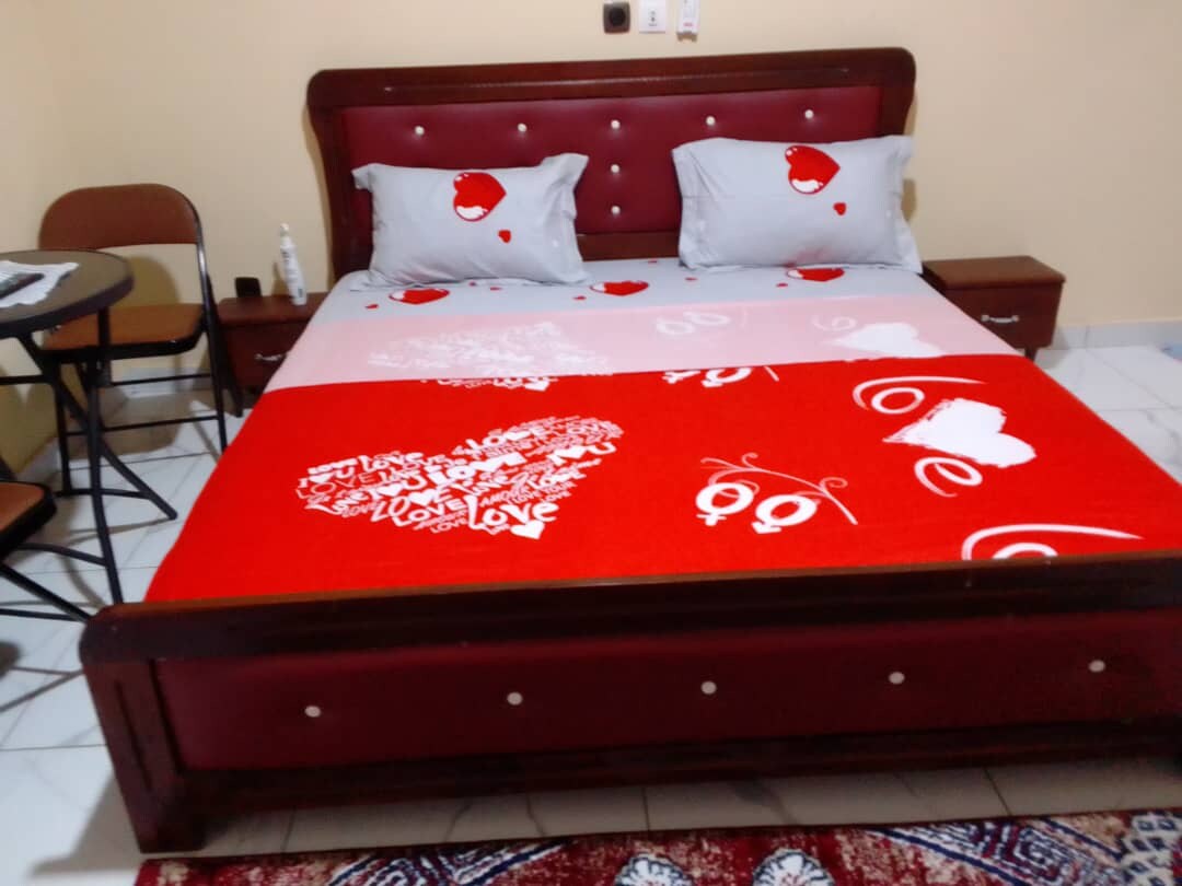 A spacious bed is adorned with a bright red and white blanket featuring heart motifs. Two decorative pillows sit against the headboard, providing comfort. A small table and chair are positioned nearby, complementing the simple and functional design of the room.