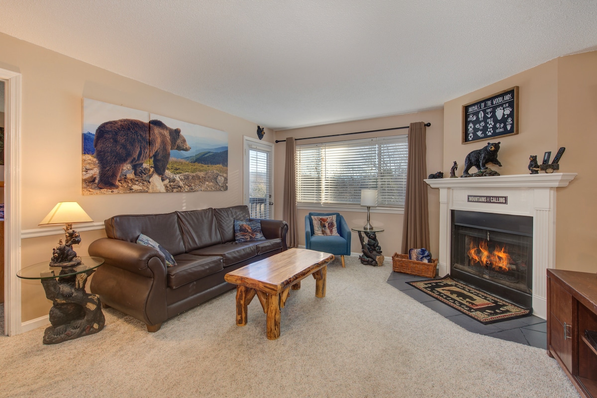 A cozy living room is highlighted by a stone fireplace and large windows allowing natural light. A leather sofa faces the fireplace, complemented by a wooden coffee table. A bear-themed artwork adorns the wall, and a comfortable chair is positioned nearby.