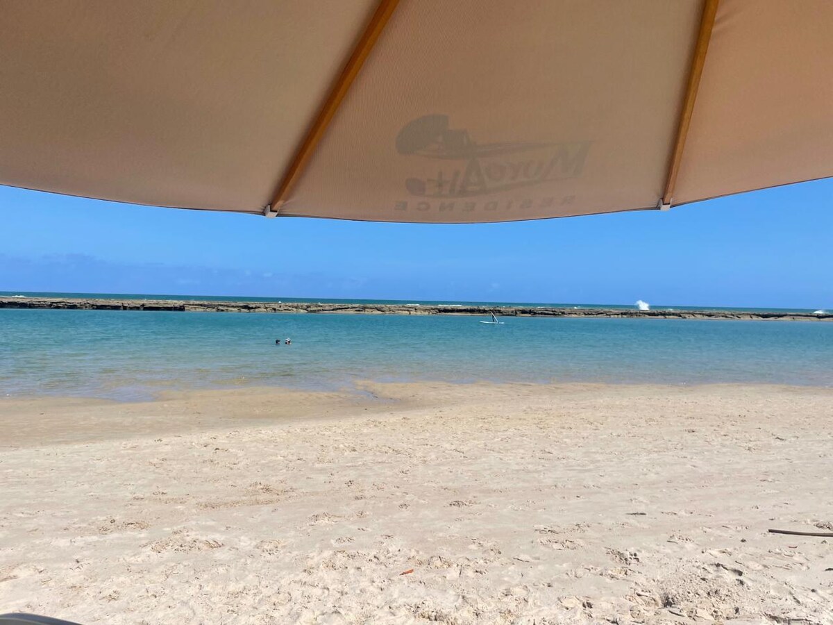 A sandy beach is seen under a large umbrella, with calm waters stretching toward the horizon. The clear blue sky complements the serene landscape, while distant rocks form a boundary in the water. Gentle waves are visible, enhancing the tranquil scene.