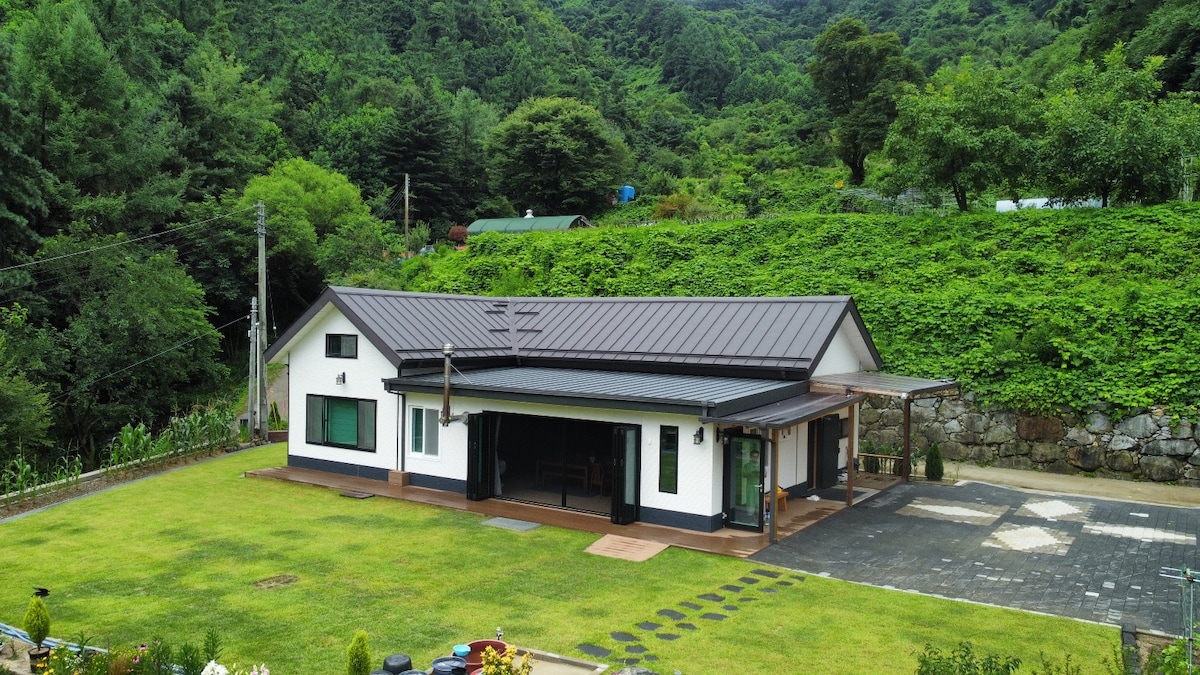 The spacious property is surrounded by lush greenery and mountains, featuring a well-maintained lawn and a patio area. The home showcases a contemporary architectural style with large windows, offering a bright and airy appearance. Ample outdoor space supports activities or relaxation.