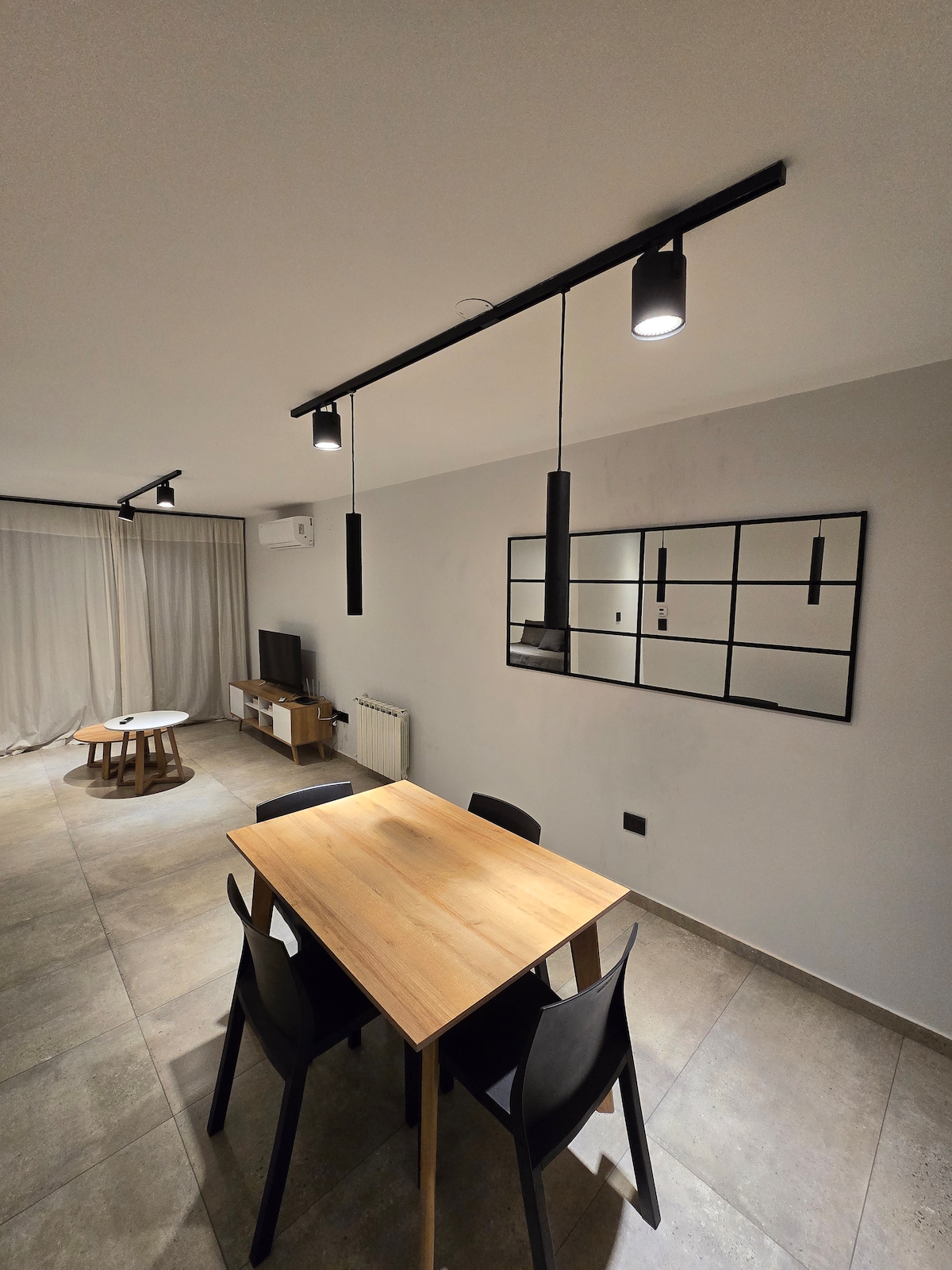 A modern dining area is presented with a wooden table surrounded by four black chairs. Above, suspended lighting fixtures provide illumination. A large mirror hangs on the wall, reflecting the space, while a cozy living area is visible in the background.