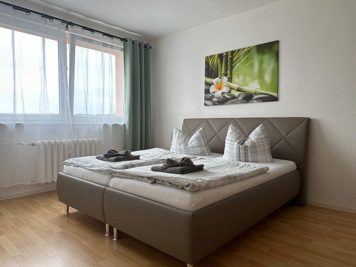 The sleeping area features a spacious bed with decorative pillows, set against a light-colored wall. A large window allows natural light to fill the room, complemented by sheer curtains. A peaceful wall art piece showcases bamboo and stones, adding a calming touch.