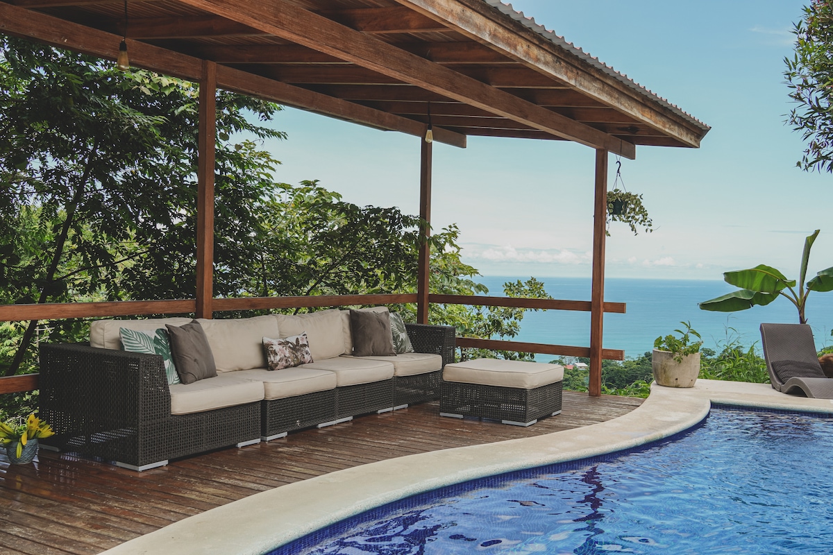 A covered terrace features a comfortable seating area with neutral cushions facing an ocean view. A smooth, curved edge of a swimming pool is positioned nearby, surrounded by greenery and overlooking the distant horizon.