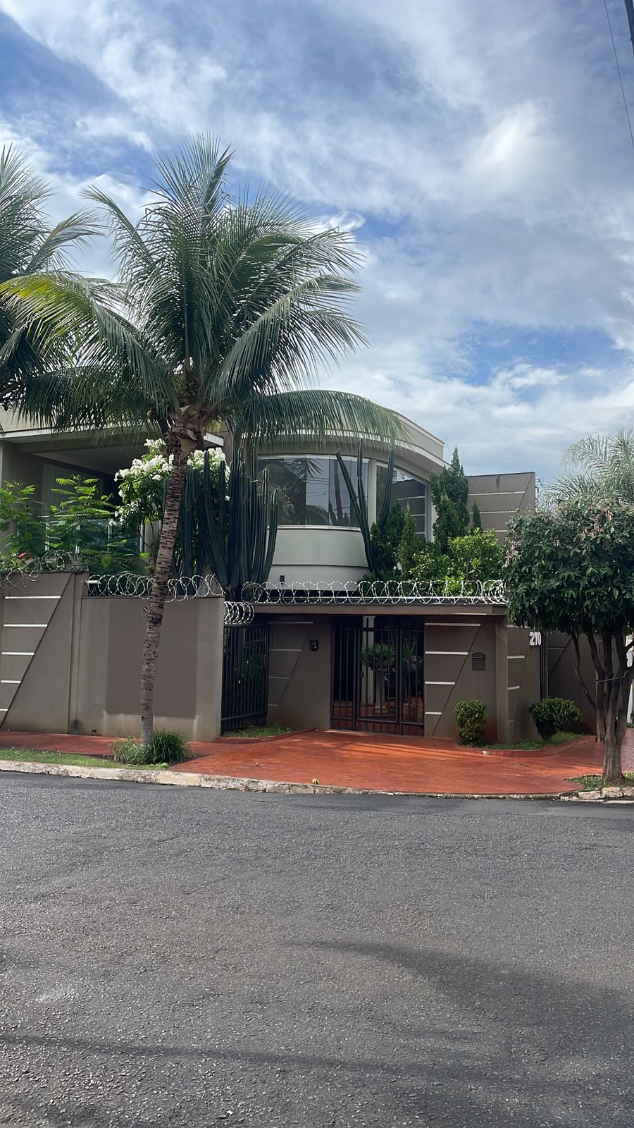 The exterior of a modern house is shown, framed by lush palm trees. A gated entrance is visible, with desert-style landscaping and a circular driveway. The building combines light-colored walls with a rounded upper level and is situated on a quiet street.