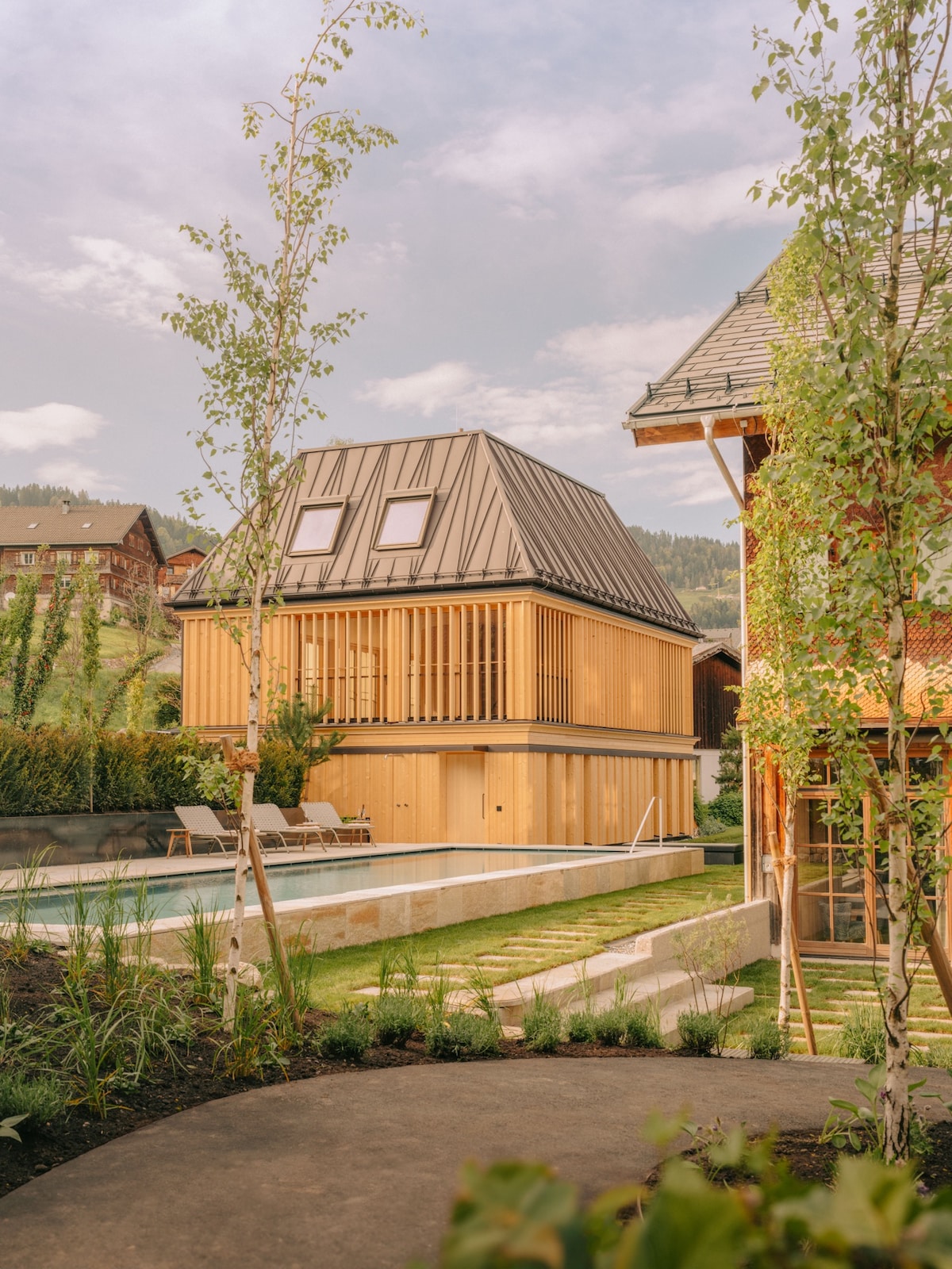 The exterior of a contemporary wooden building is captured, featuring a sloped roof and large, horizontal wooden slats. A tranquil swimming pool is positioned in front, surrounded by well-maintained green grass and lush plants. The setting offers a serene view of the surrounding hills.