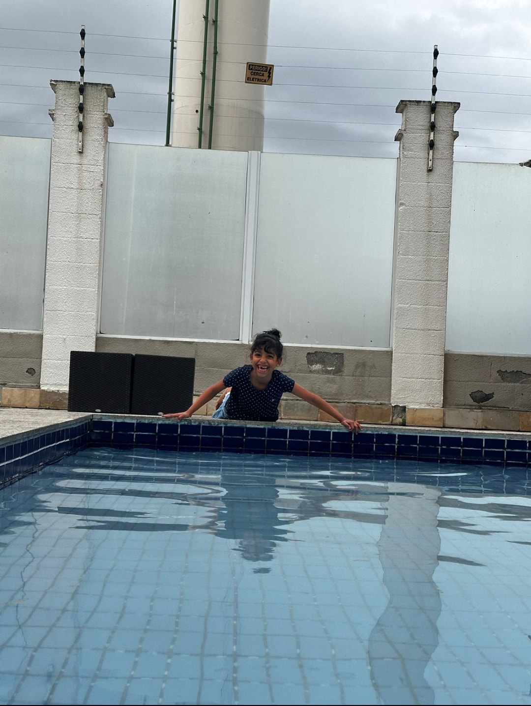 A clear swimming pool is visible, featuring blue tiles and a calm water surface. The pool is framed by white walls, with two tall columns positioned on either side, providing a sense of enclosure. The area surrounding the pool offers ample space for relaxation.
