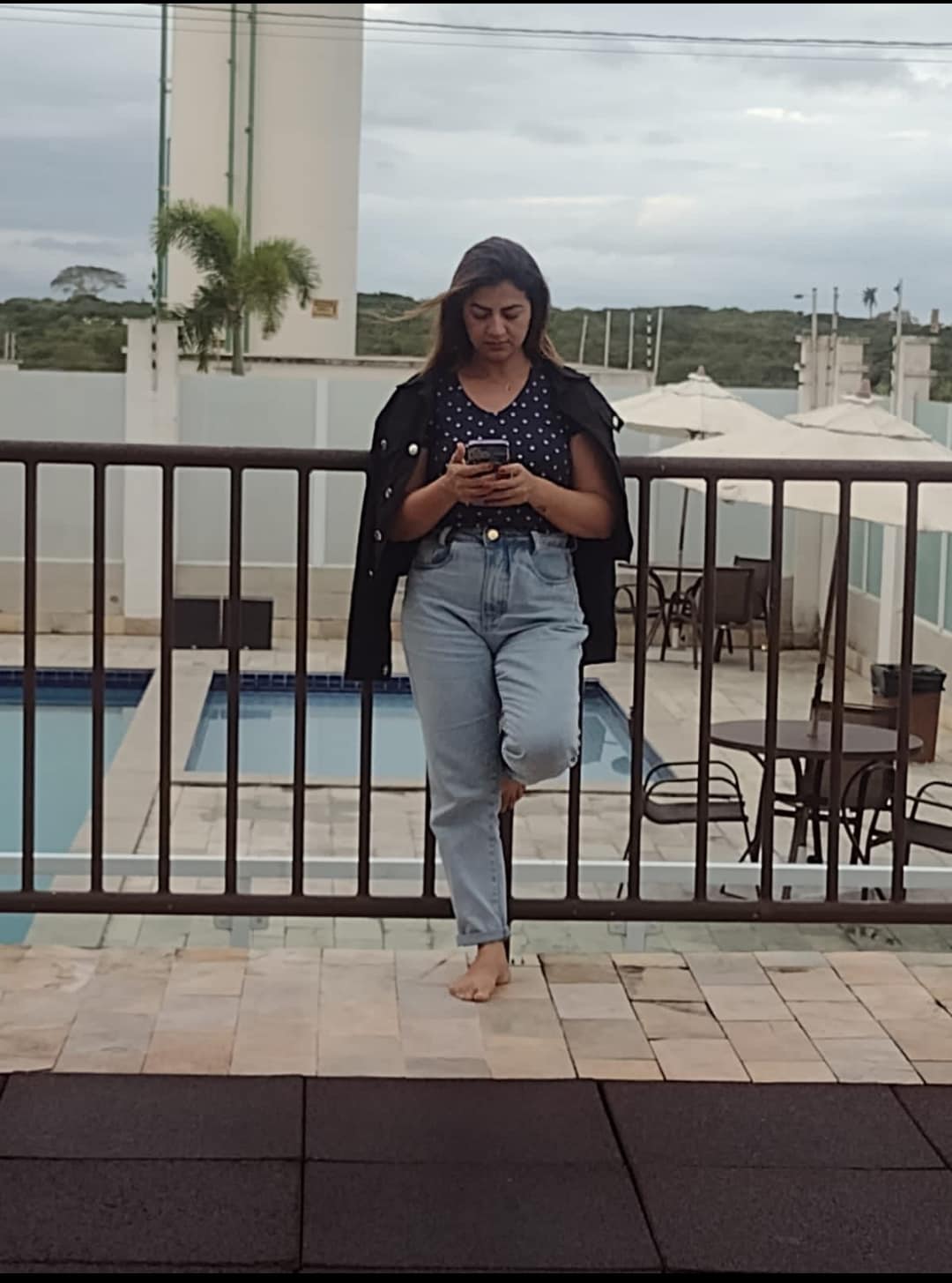 A person stands casually by a balcony railing, looking at their phone. In the background, a well-maintained pool is visible, surrounded by lounge chairs and shaded umbrellas, enhancing the relaxing environment.