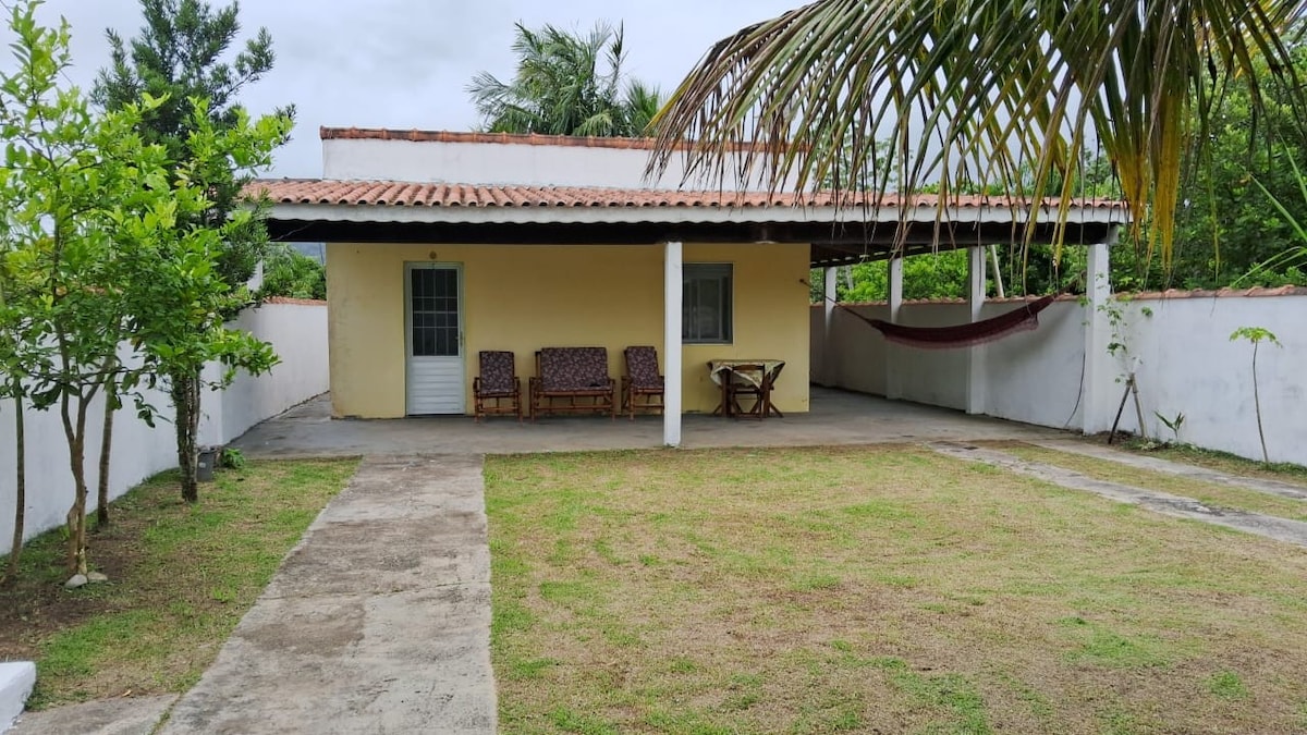 A single-story house is presented with a yellow exterior and a tiled roof. The outdoor area features a well-manicured grassy lawn, a seating arrangement of chairs, and a small table. A hammock is easily accessible, and tropical plants are positioned along the perimeter.