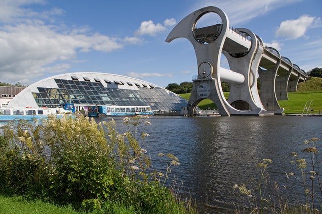 The image showcases the impressive architecture of the Falkirk Wheel, a unique boat lift, alongside the visitor center. The structure's distinctive curves and angles are contrasted by the calm waters of the surrounding canal and lush green landscaping, under a clear blue sky.