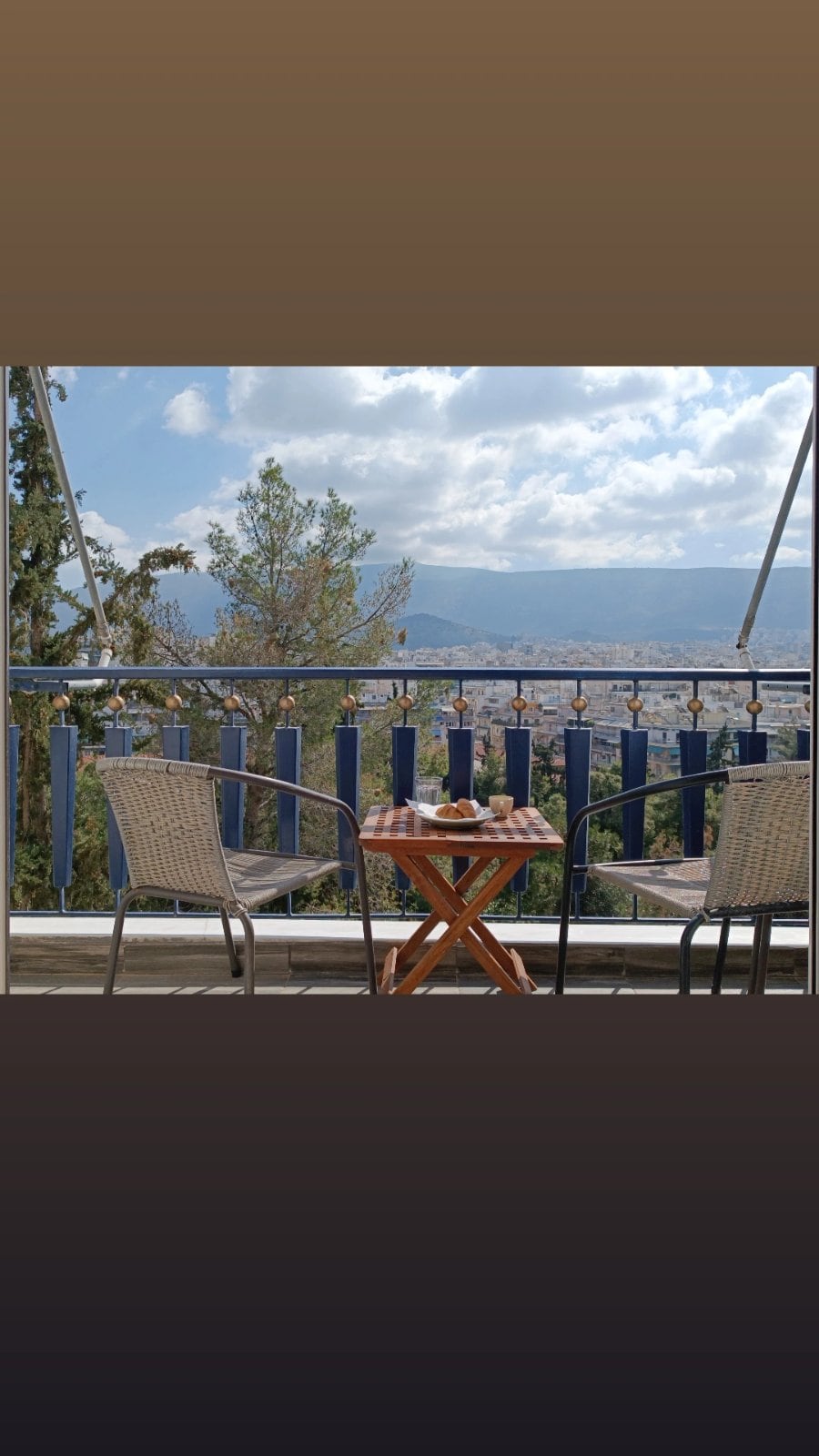 A charming balcony features two wicker chairs flanking a small wooden table. A picturesque view of the surrounding greenery and distant mountains is visible in the background, providing a serene space for relaxation.