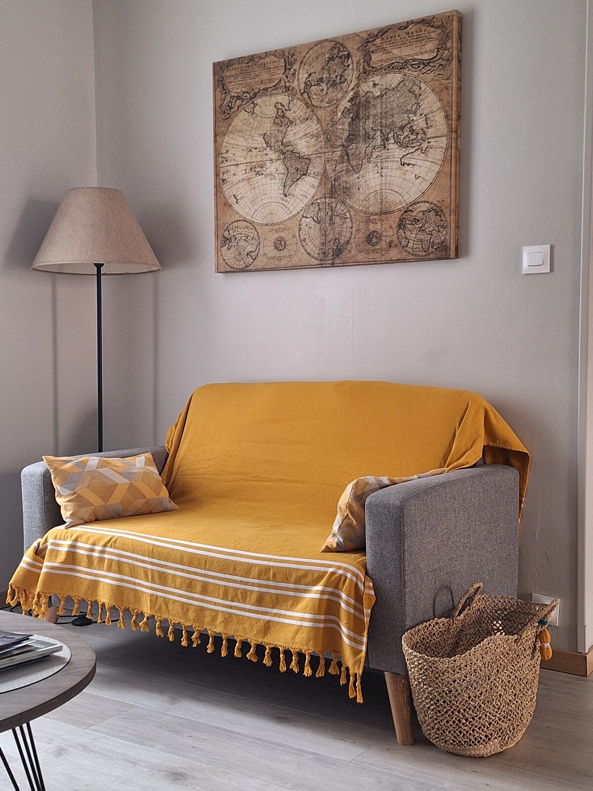 A cozy seating area is featured, with a gray couch draped in a yellow throw. A small patterned cushion adds a touch of color. A vintage map artwork adorns the wall above. A woven basket sits beside the couch, contributing to the relaxed ambiance.