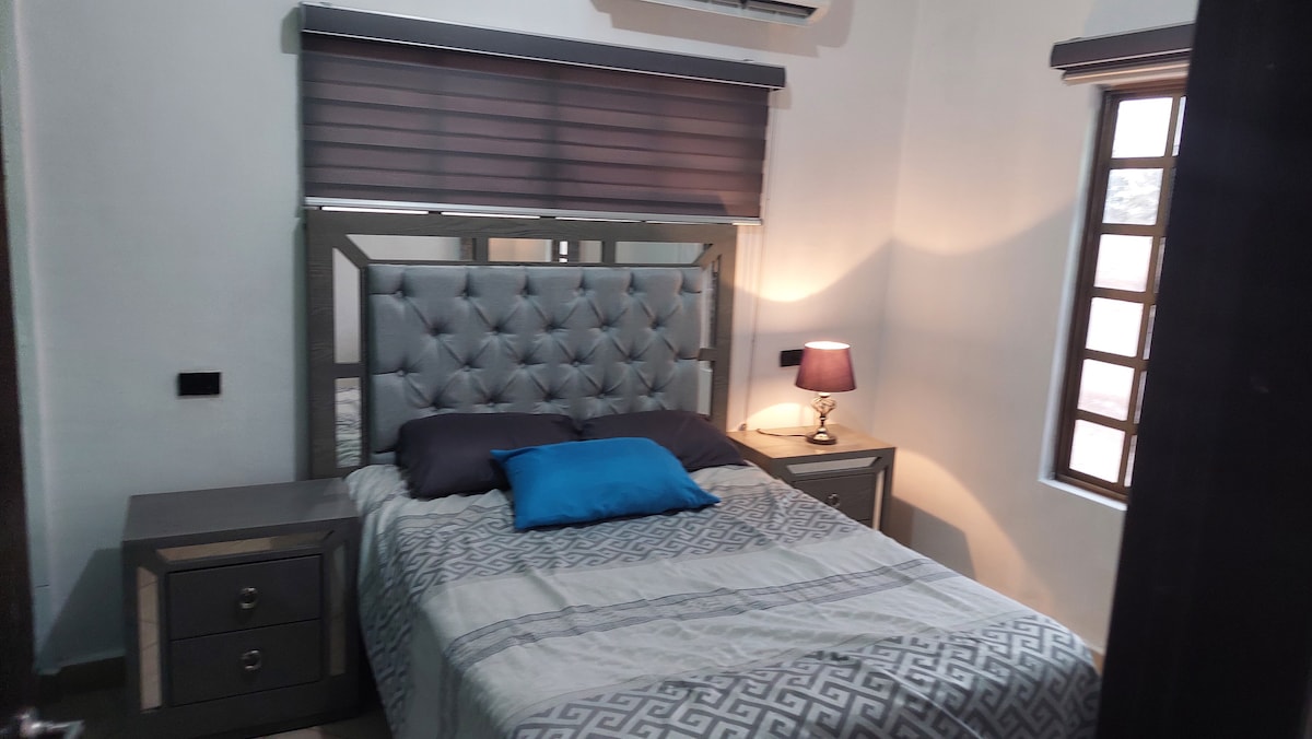 A cozy bedroom is featured, highlighted by a tufted headboard and a colorful accent pillow on a spacious bed. A bedside table with a lamp stands beside the bed, and natural light enters through a window covered by horizontal blinds.