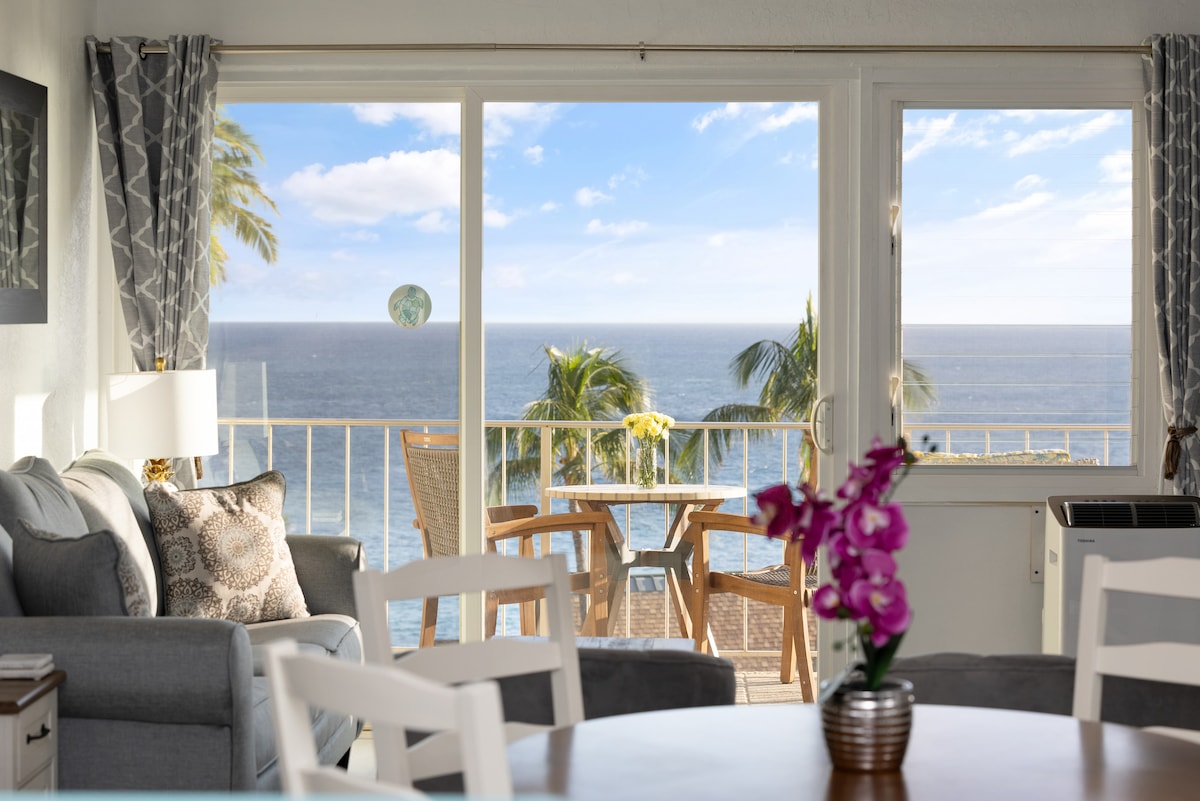 A spacious living area is presented, featuring a comfortable grey sofa and a dining table. A large window offers ocean views, framed by light curtains. A small outdoor balcony with seating is visible, complemented by tropical palms and blue skies outside.