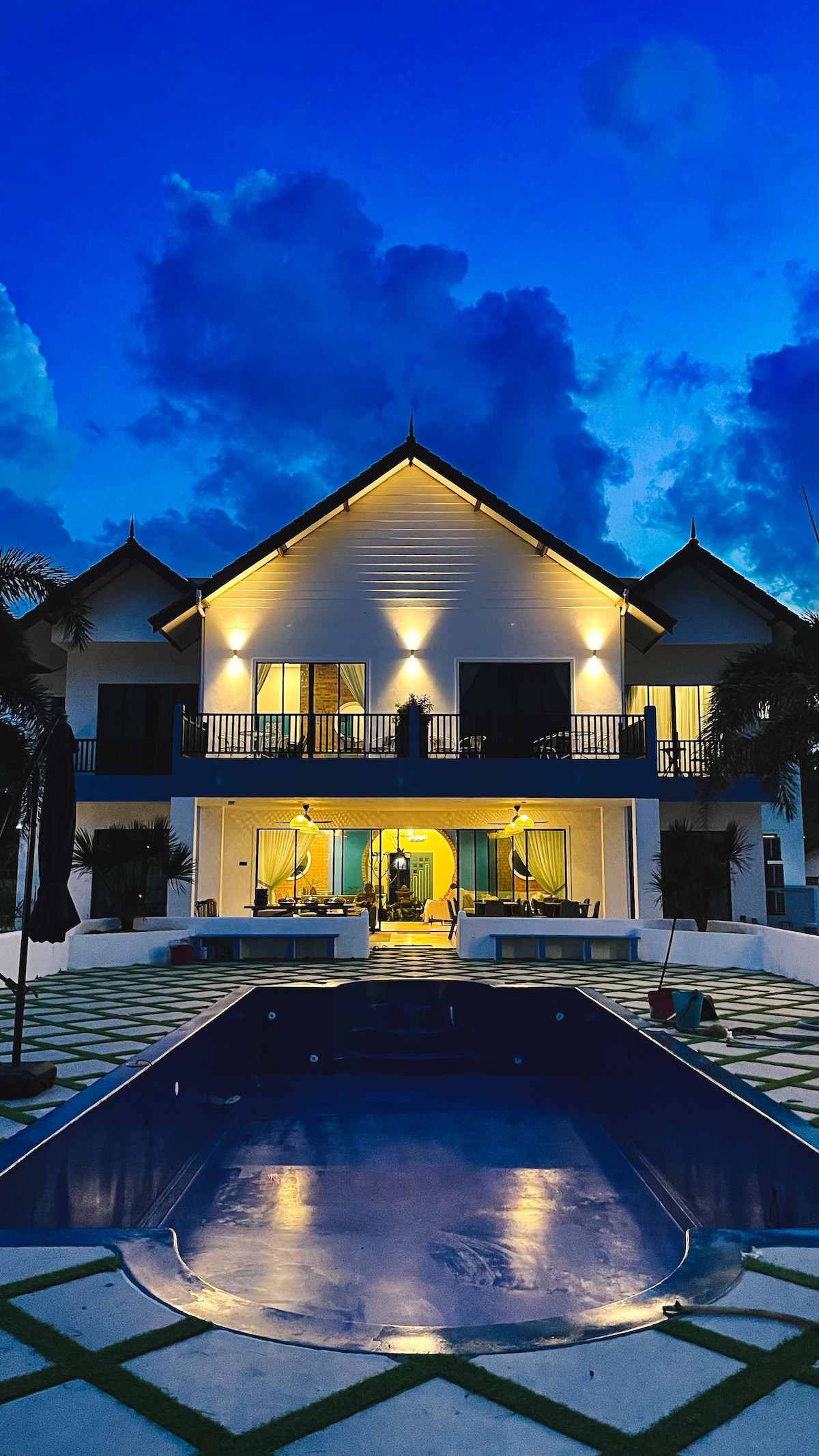A two-story villa is illuminated against a twilight sky, featuring balconies and lush landscaping. The pool in the foreground reflects the evening light, inviting relaxation. Fluffy clouds create a dramatic background, enhancing the overall serene atmosphere of the property.