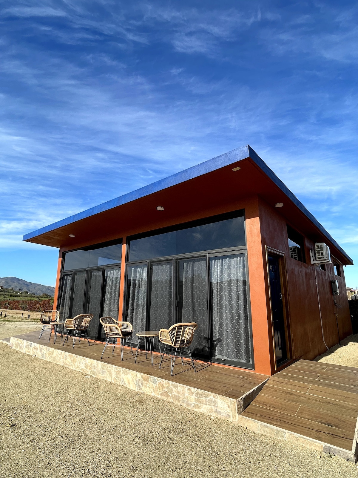 A modern casita exterior is highlighted by large glass windows and a sloped roof. A small deck with four woven chairs and a table is visible in front, offering a view of the surrounding rural landscape, complemented by a clear blue sky.