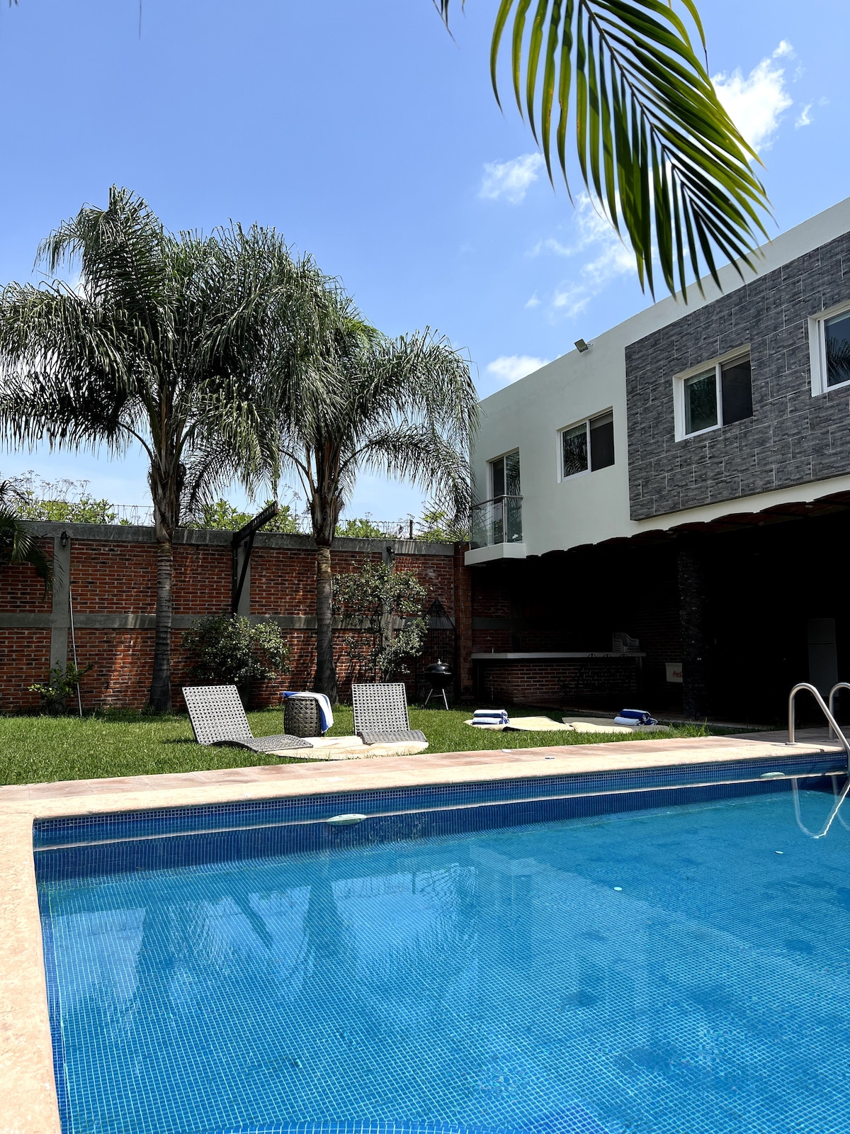 A spacious outdoor area is showcased with a well-maintained pool, surrounded by lounge chairs and tropical palm trees. A brick wall provides privacy, while part of the house is visible in the background, displaying a modern design and large windows.
