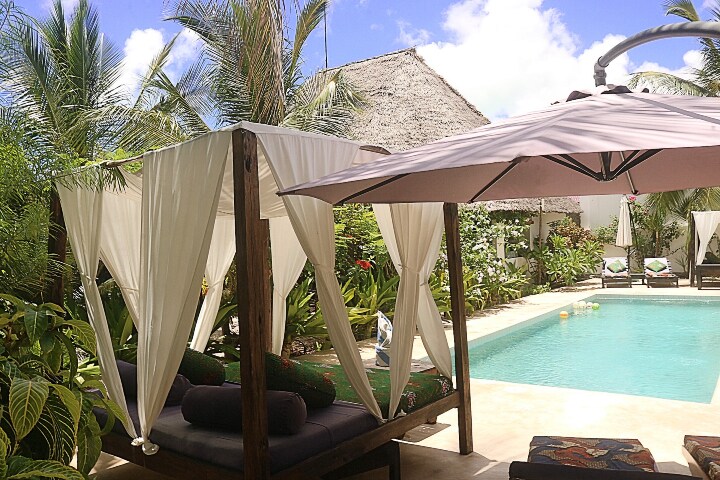 A tranquil lounge area features elegantly draped canopies surrounding comfortable seating. A large umbrella provides shade over the space, while a serene swimming pool is framed by lush tropical plants and palm trees, contributing to a peaceful outdoor setting.