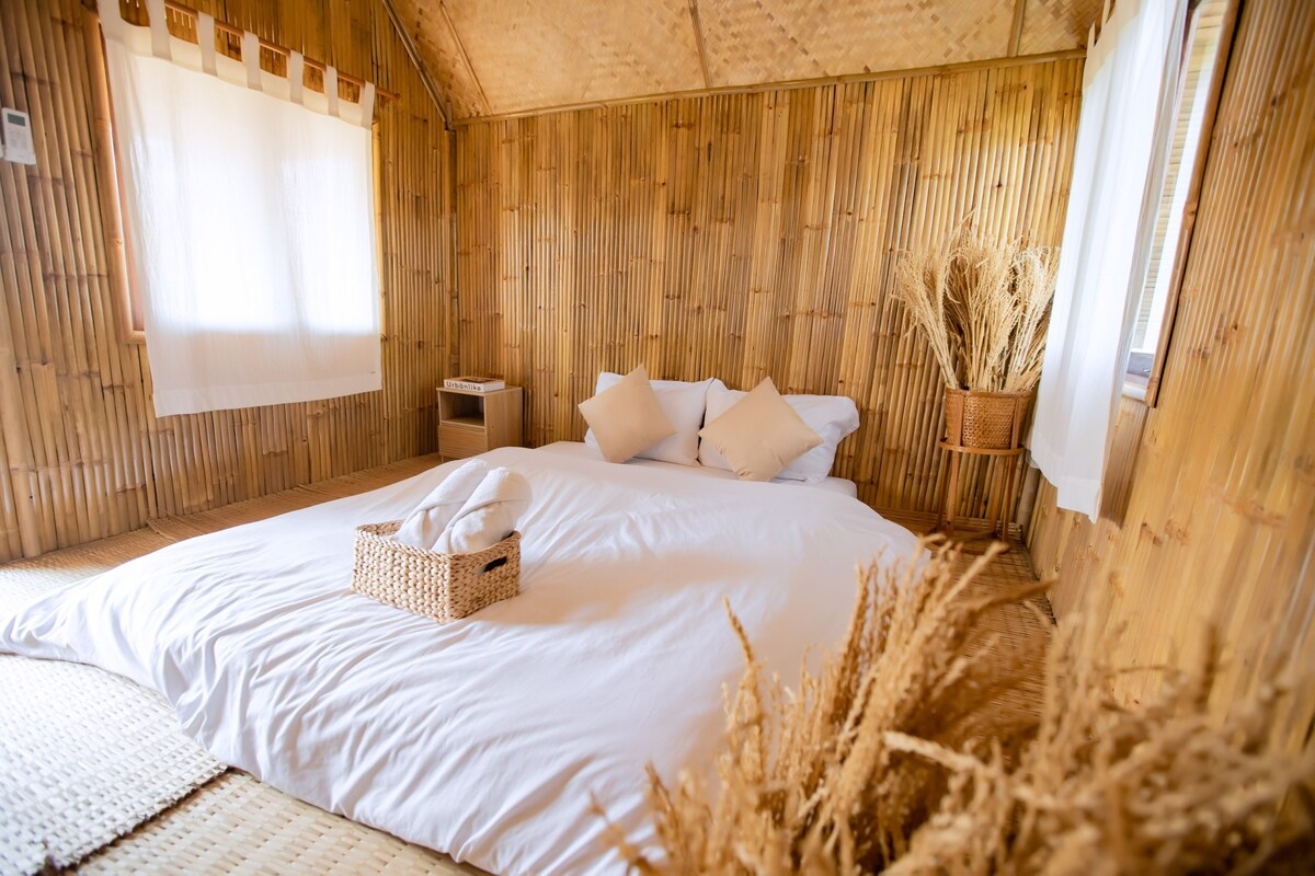 The interior features bamboo walls and a spacious bed adorned with white linens and decorative pillows. Natural light filters through sheer white curtains, highlighting a woven basket and dried plants in the corner, creating a serene environment.