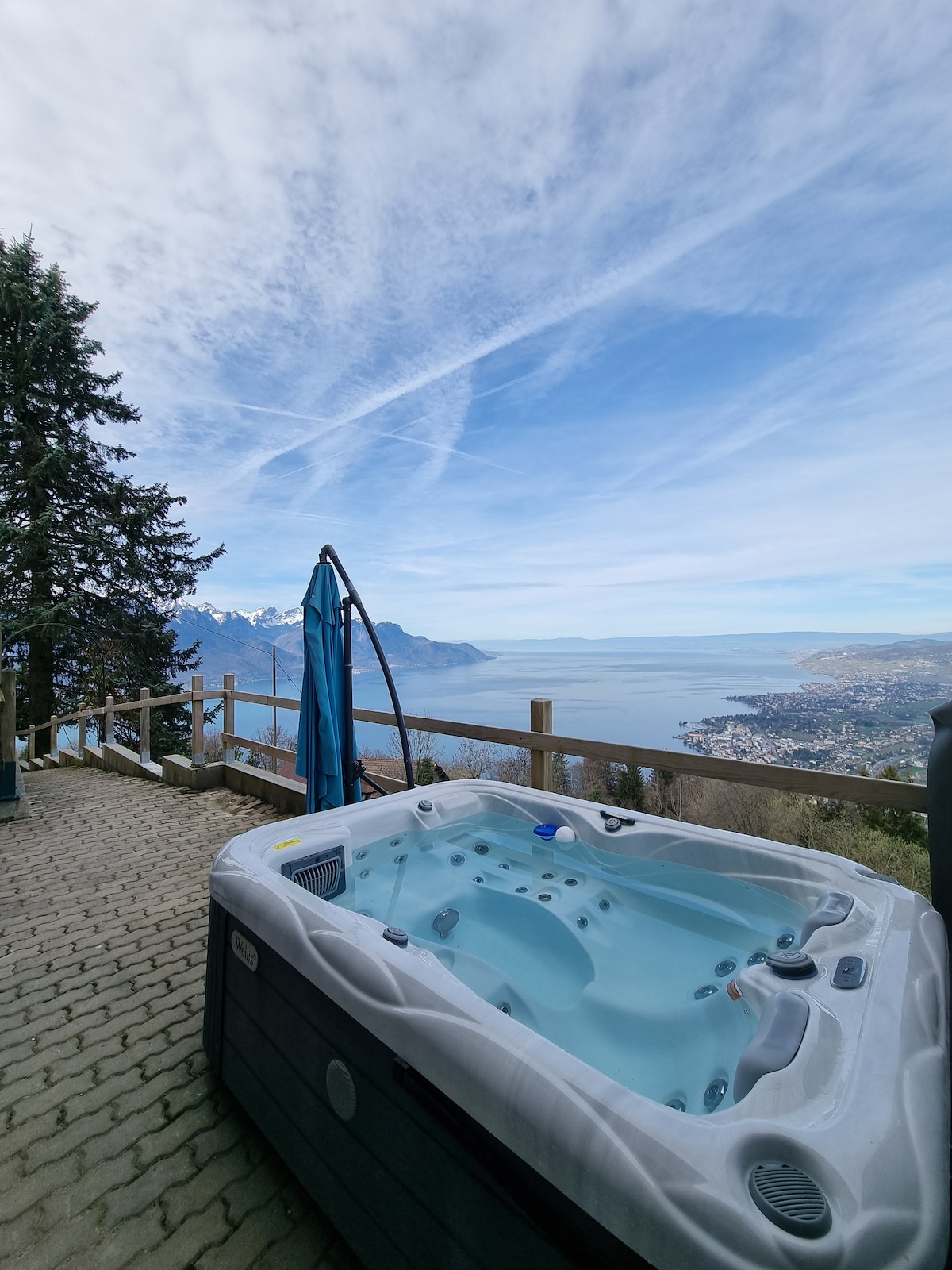A spacious outdoor jacuzzi is set on a deck, offering breathtaking views of a lake and nearby mountains. Tall trees surround the area, enhancing the natural setting. A sunshade is positioned beside the jacuzzi for added comfort.