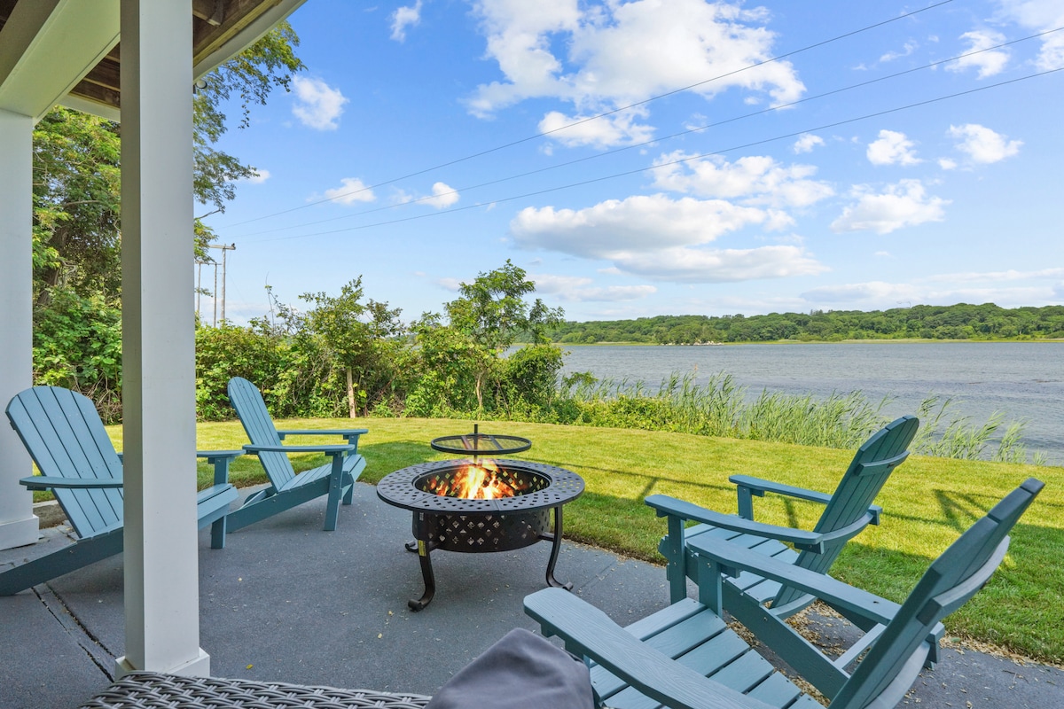 An outdoor patio area is visible, featuring several teal Adirondack chairs arranged around a central fire pit. Lush greenery borders the space, overlooking a tranquil water view under a clear blue sky with scattered clouds.