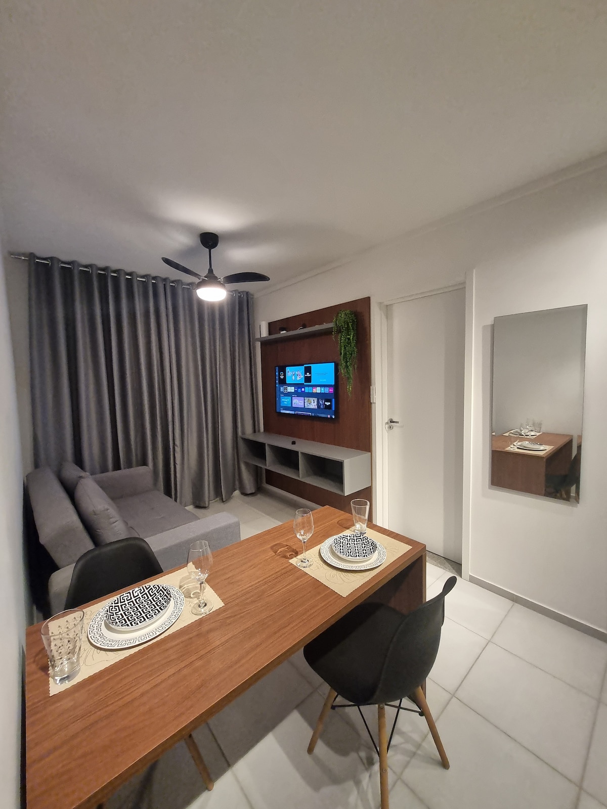 The dining area features a wooden table set for two, with elegant table settings. A comfortable gray sofa is positioned against a wall, accompanied by a ceiling fan above. A mounted television is visible on the adjacent media console, complemented by indoor greenery and a mirror reflecting the space.