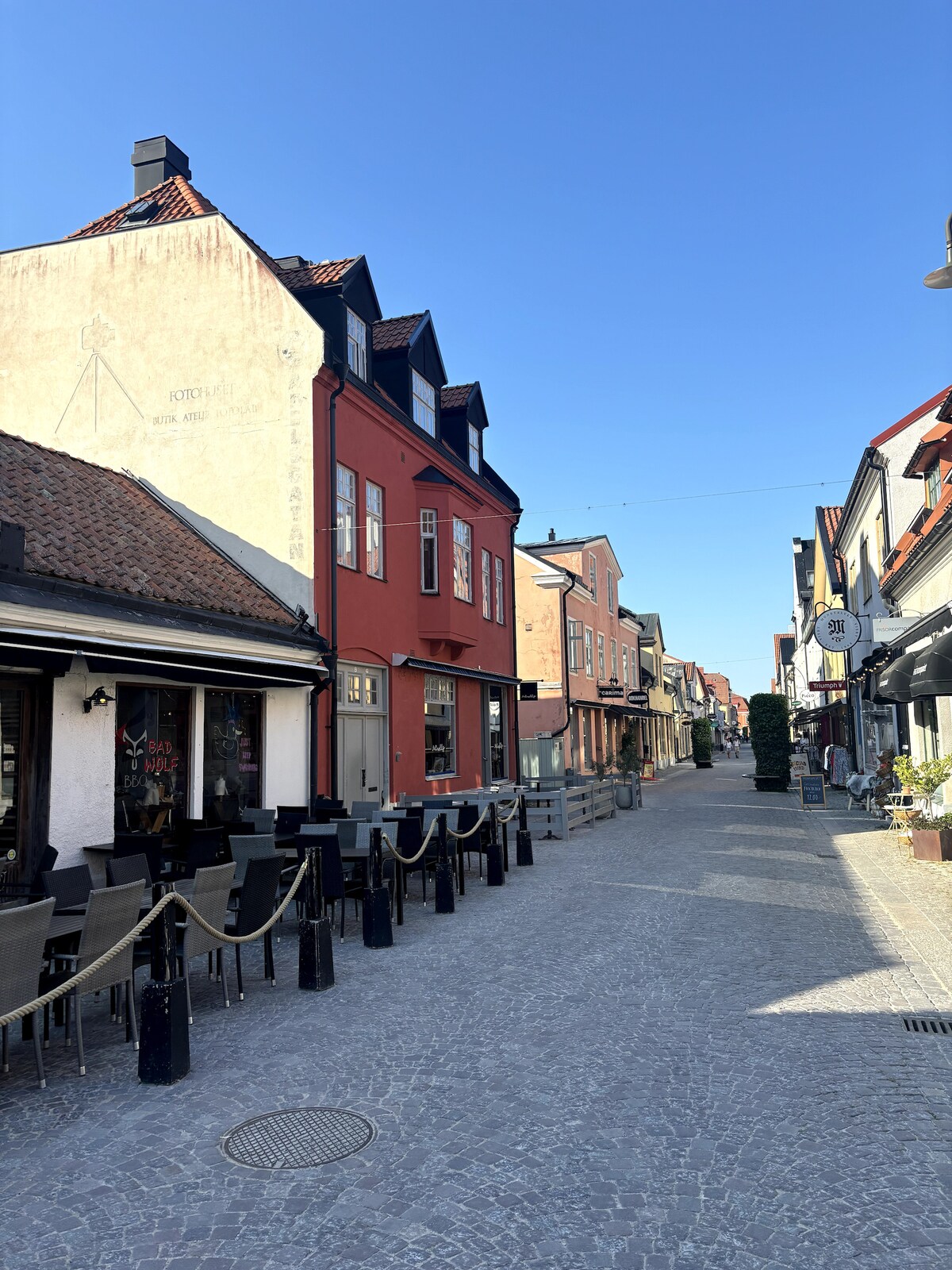 A cobblestone street is lined with charming buildings featuring a mix of red and traditional architecture. Outdoor seating is visible along the sidewalk, creating a welcoming atmosphere. Bright blue skies are present, enhancing the inviting ambiance of the area.