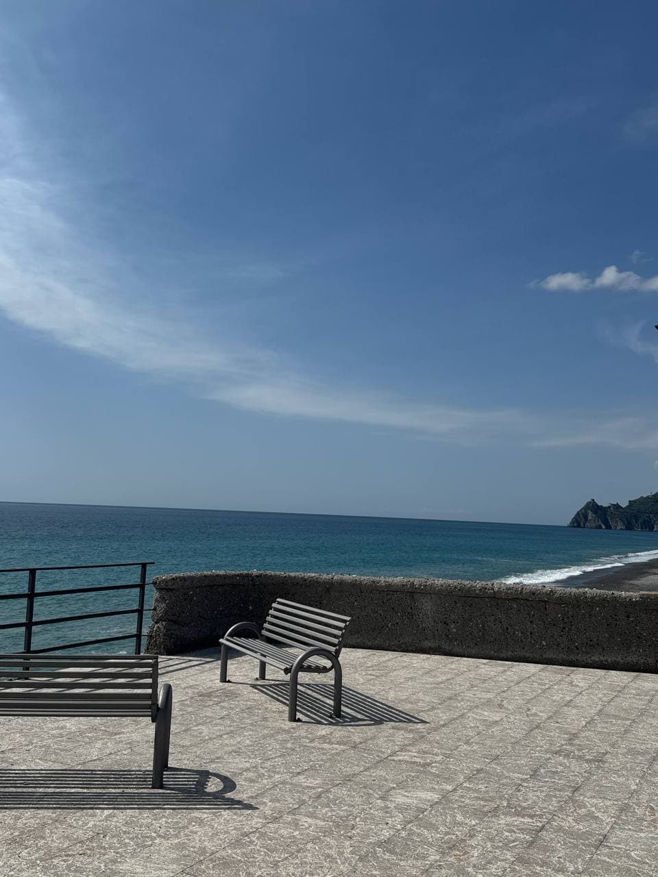 An outdoor area features a stone pathway leading to a pebbled beach. Two metal benches are positioned along the edge, facing a calm sea under a clear blue sky. Gentle waves lap the shore, and a distant rocky coastline is visible.