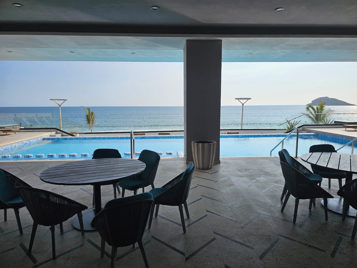 An outdoor area features a spacious swimming pool surrounded by lounging spaces. Various chairs are arranged around round tables, offering sights of the ocean through large openings. Sunlight brightens the area, highlighting the pool's calm waters and the vista of the beach in the background.