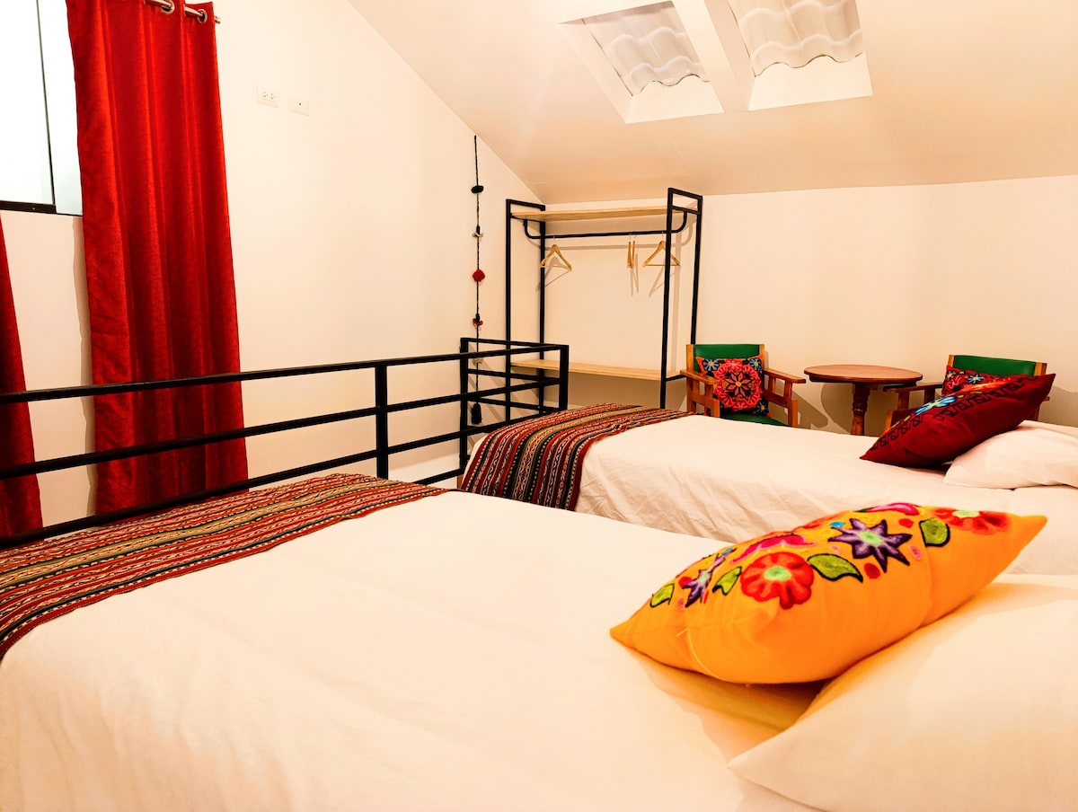 The room features two spacious beds adorned with bright, decorative pillows. A small round table and two colorful chairs are positioned nearby. A clothing rack is visible, and light enters through a window covered by red curtains.