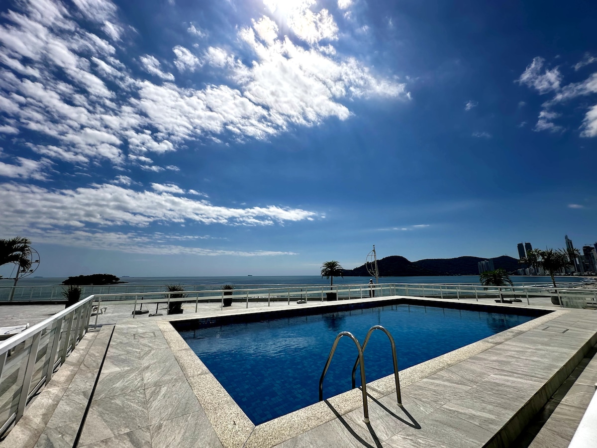 A swimming pool is set against a backdrop of the ocean and sky, featuring clear water and a surrounding sunlit deck. Palm trees are visible in the area, and distant mountains enhance the scenic view. The atmosphere appears calm and inviting.