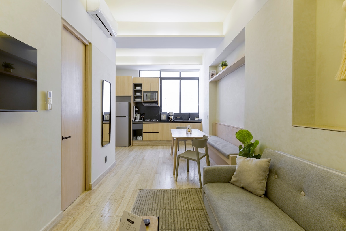 A modern living area features a light-colored sofa alongside a minimalist dining table. The fully equipped kitchen with appliances is visible in the background, complemented by large windows that allow natural light to fill the space, creating a bright and airy atmosphere.
