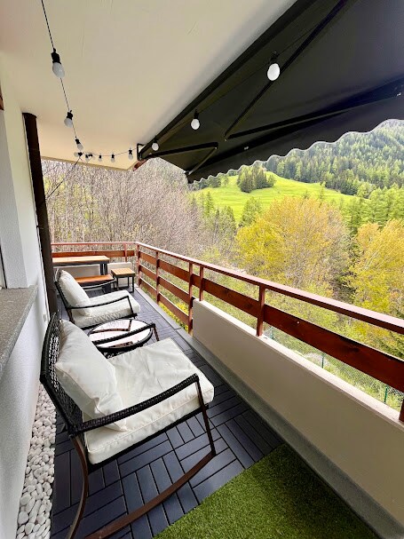 A cozy balcony is furnished with two cushioned armchairs and a small table. String lights are hung overhead, creating a gentle ambiance. The view showcases a lush green landscape, framed by trees, providing a serene outdoor escape.