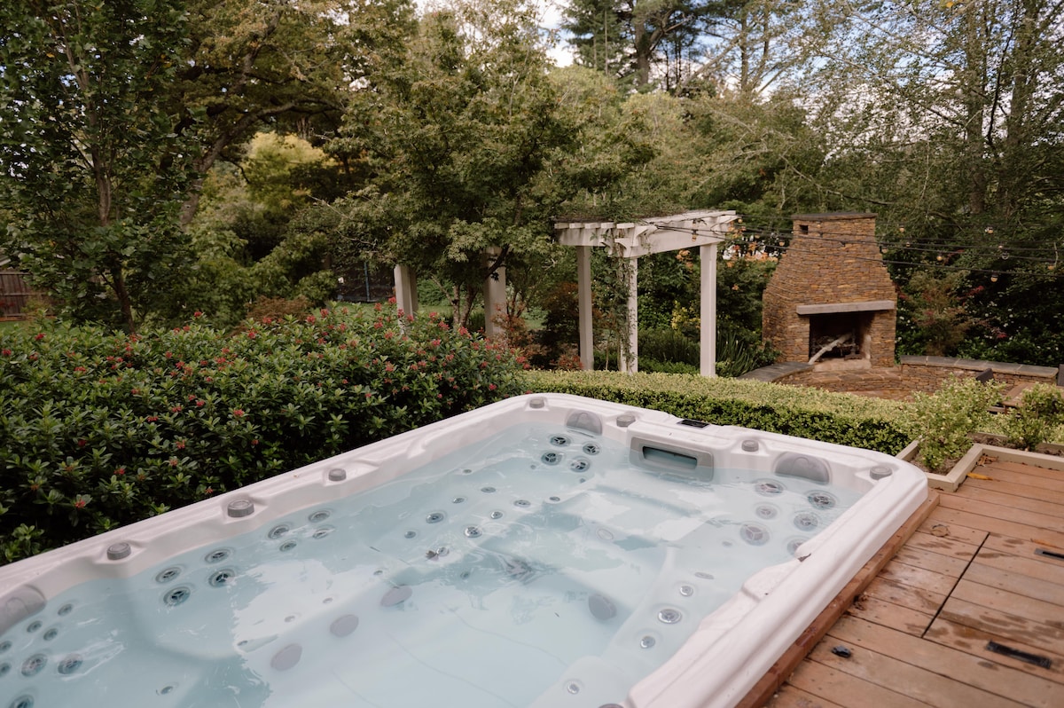 An outdoor spa is set on a wooden deck, surrounded by lush greenery and flowering shrubs. Nearby, a stone fireplace is nestled within the landscape, adding a cozy element to the tranquil garden setting.