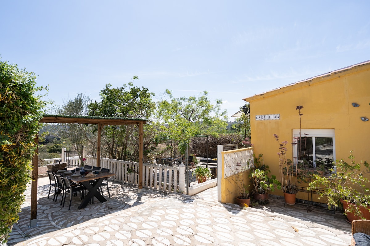 A sunlit patio area is visible, featuring a large dining table under a wooden pergola. The surrounding space is decorated with plants and provides a view of the lush garden and hills beyond. The yellow exterior of the adjacent house adds a welcoming touch to the setting.