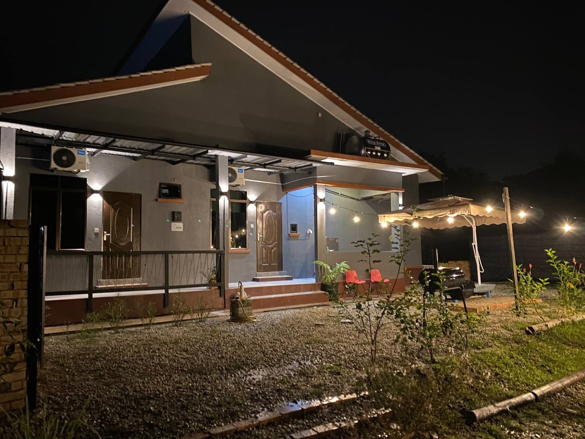 The exterior of the property is illuminated at night, showcasing a spacious front area with gravel pathways. A covered seating area with chairs and a table is visible, accompanied by string lights that add a gentle glow. Nearby, lush greenery enhances the secluded ambiance.