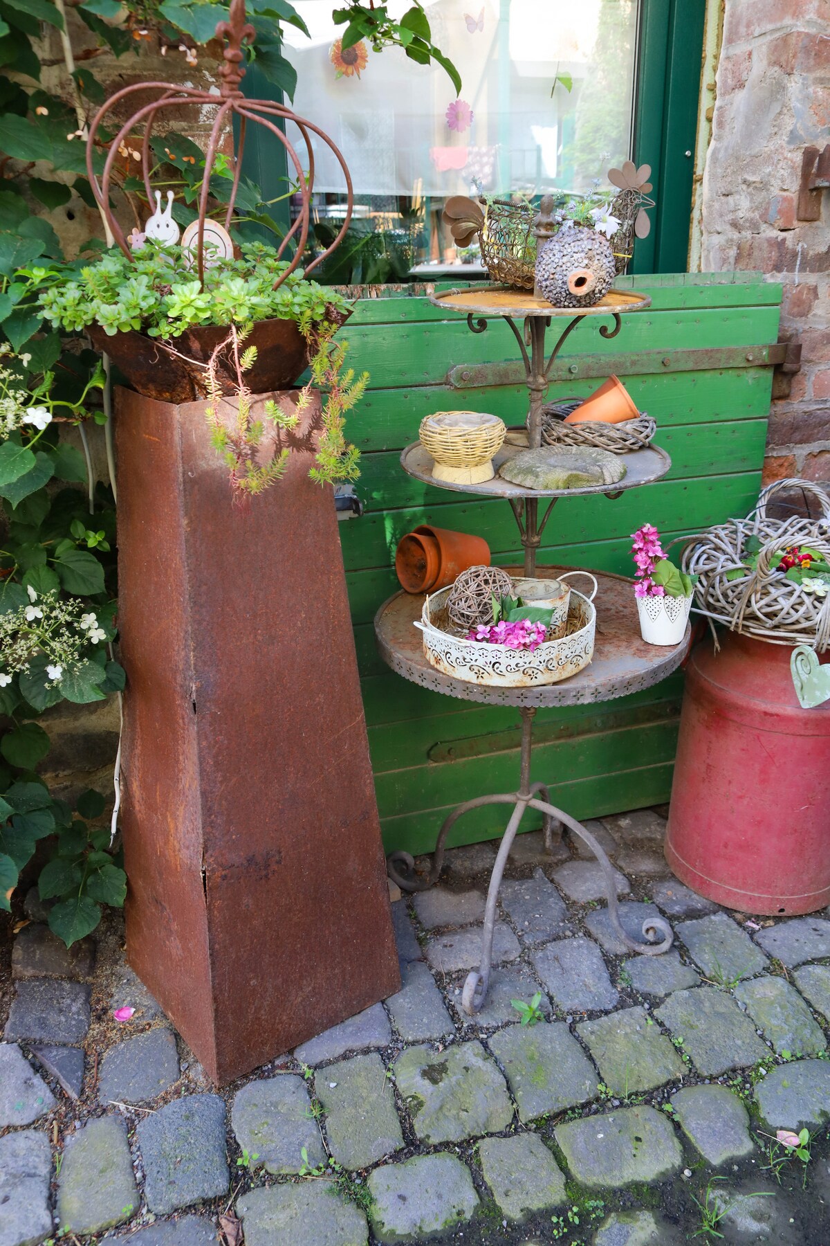 An arrangement of decorative items and plants is displayed on a metal stand beside a weathered green wall. A rusted planter holds lush greenery, while various baskets and pots are artistically arranged on the stand, adding a charming touch to the outdoor space.