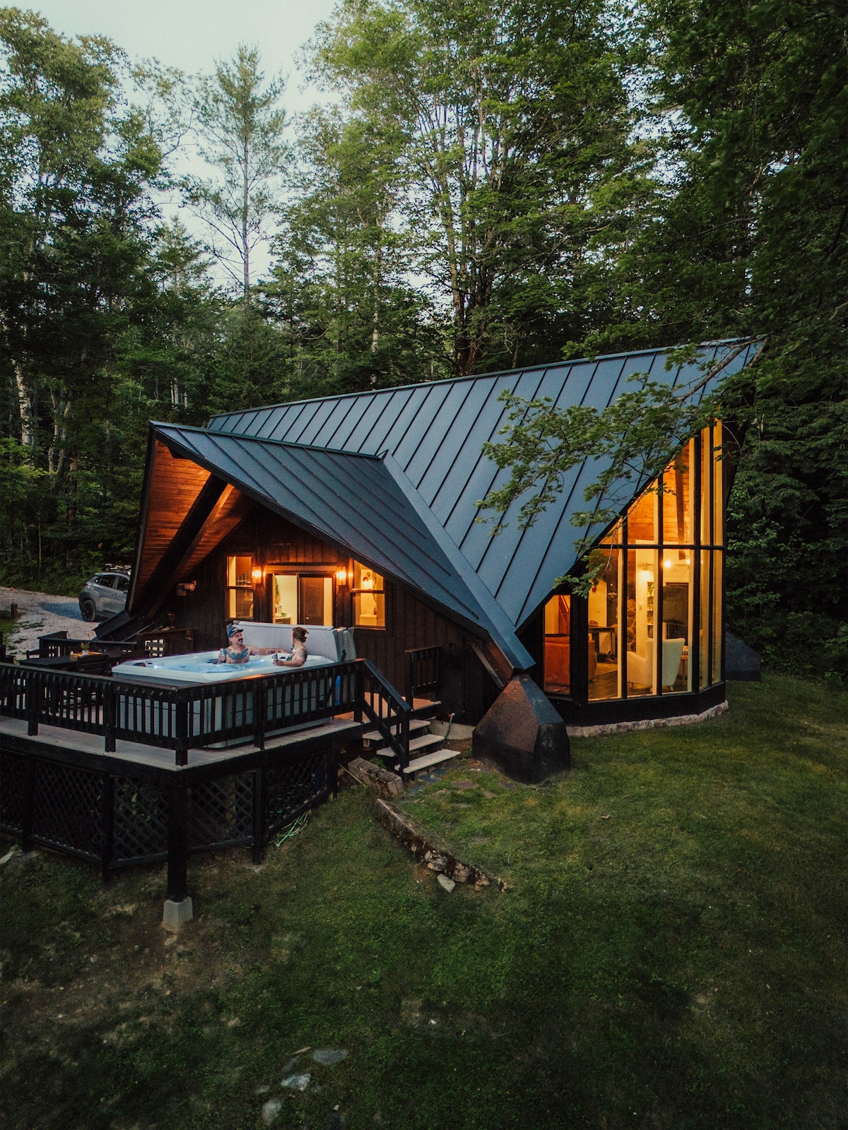 An A-frame cabin with a distinctive angled roof is set amidst lush greenery. The exterior features large windows that illuminate the interior, while a spacious deck is equipped with seating. The tranquil surroundings create a serene atmosphere for relaxation.