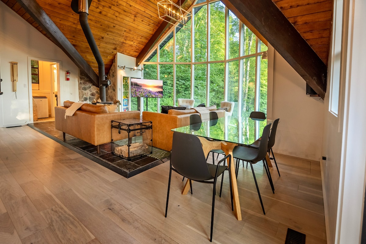 A spacious living area features a wall of windows that floods the room with natural light and offers views of the surrounding trees. Comfortable seating arrangements include a sofa and armchairs, paired with a modern dining table. A wood-burning stove is visible in the corner.