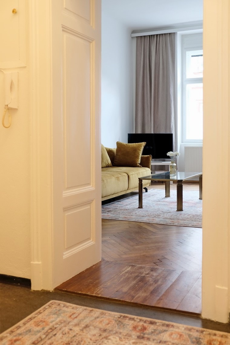 A glimpse into the spacious living area is provided through the open doorway. A comfortable sofa in a warm tone is visible, complemented by a glass coffee table on an area rug. Natural light enters through a large window, enhancing the room's inviting appearance.