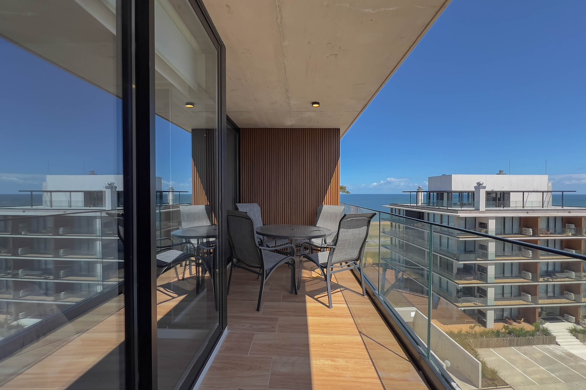 A spacious private terrace features a table with four chairs, surrounded by glass railings. The view includes nearby buildings and the ocean in the distance, with clear blue skies enhancing the outdoor space. Natural light illuminates the terrace, providing a serene spot for relaxation.