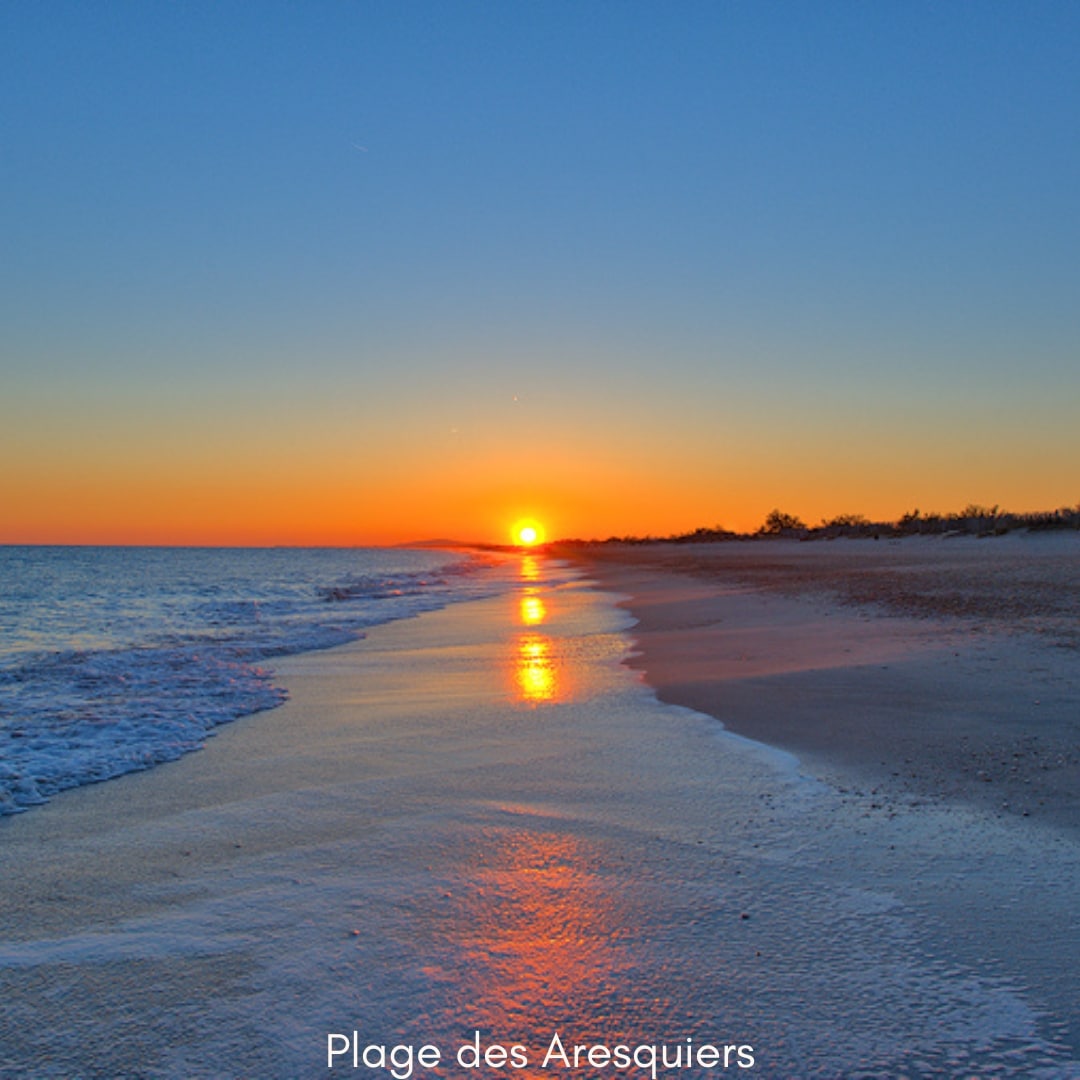 A serene beach scene captures the sun setting over the horizon, casting warm hues across the sky. Gentle waves lap at the sandy shore, reflecting the orange and blue tones of the evening light. The tranquil atmosphere suggests a peaceful moment by the coast.