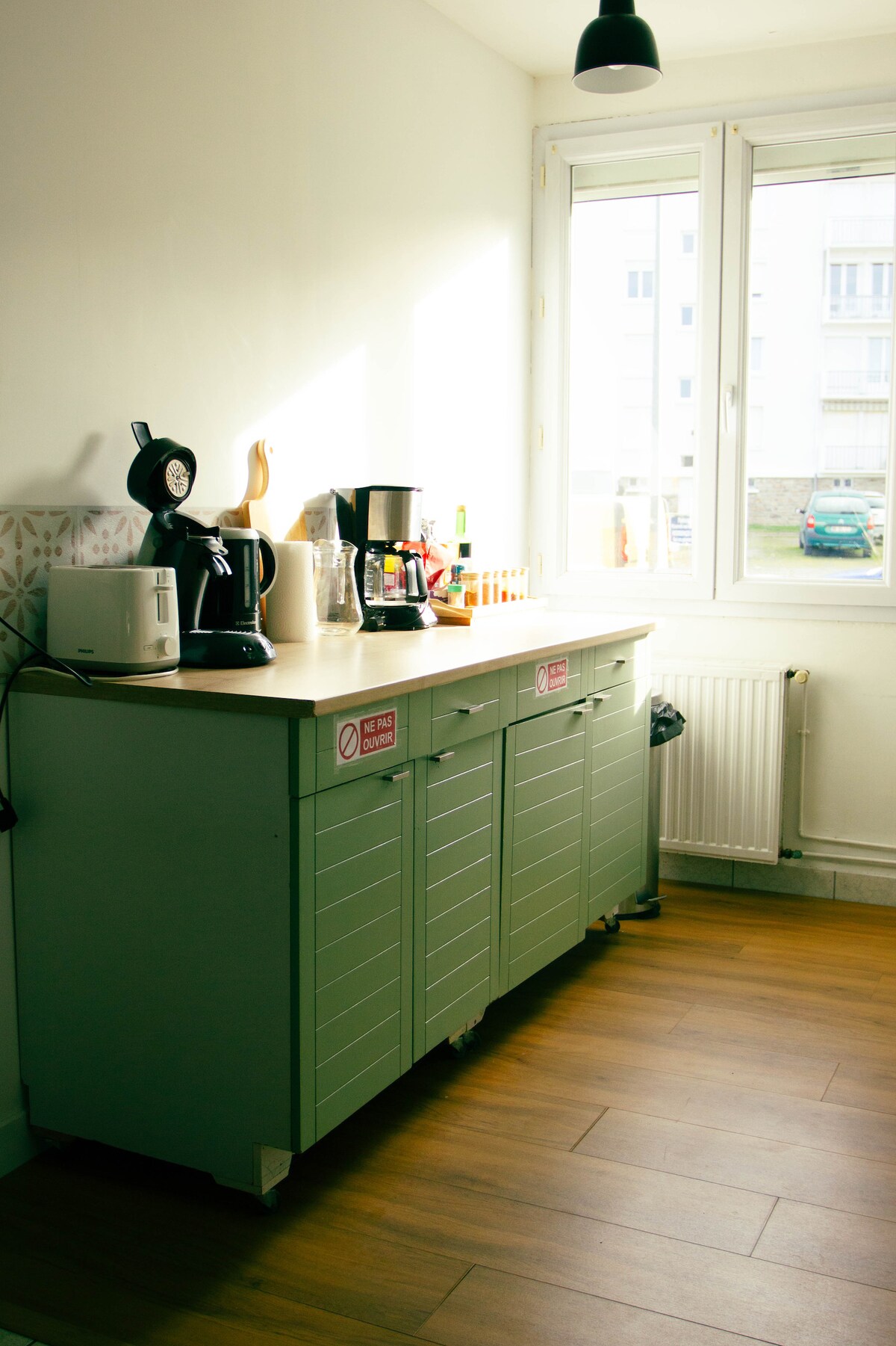 A well-organized kitchen corner features a wooden counter with a coffee maker, kettle, and additional kitchen essentials. Light filters through two windows, illuminating the space, while green cabinets below provide storage. A glimpse of outdoor greenery is visible through the windows.