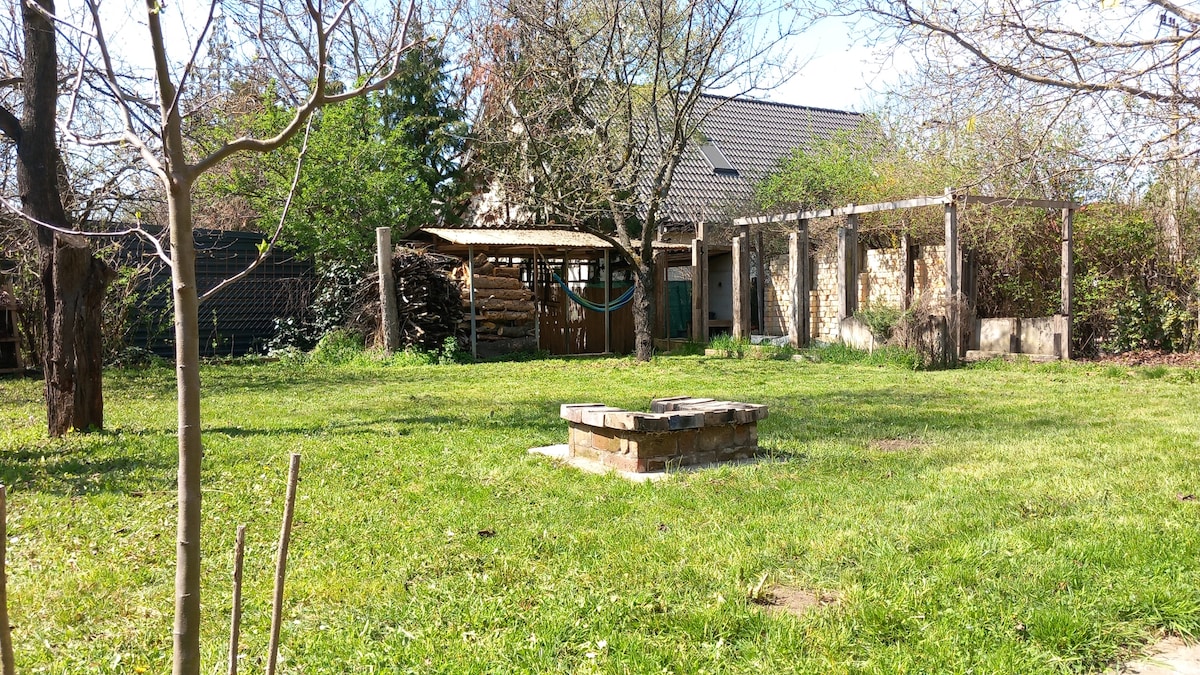 A spacious garden features a well-maintained lawn surrounded by trees. A stone fire pit is positioned at the center, while a wooden pergola with climbing plants provides shade. A separate structure can be seen to the side, offering additional outdoor space.