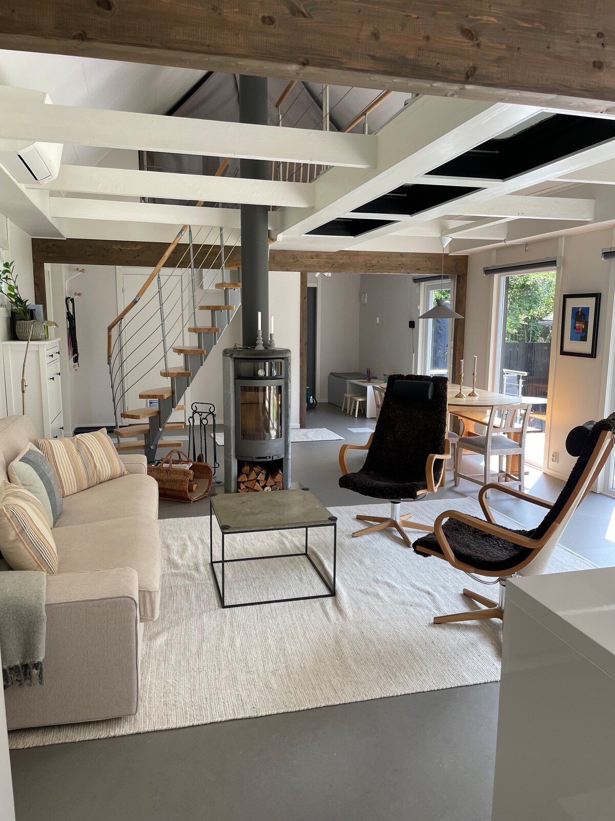 An open-plan living area features comfortable seating, including a sofa and two armchairs. A modern fireplace is positioned in the center, with a decorative frame around it. Light filters in through large windows, illuminating the space, complemented by wooden beams overhead.