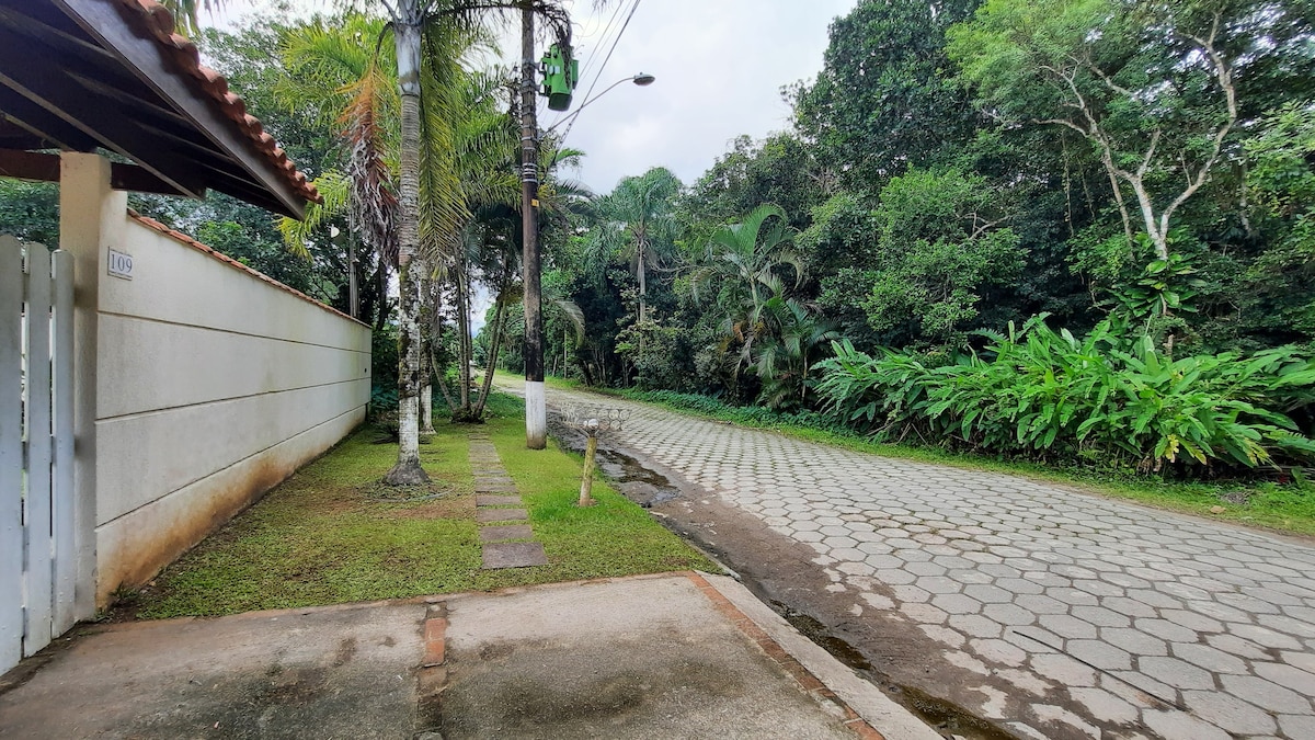 A peaceful exterior view showcases a quiet street lined with lush greenery and tropical plants. The paved road is framed by tall trees, while a well-maintained pathway leads to a white fence. The property presents a serene natural atmosphere.