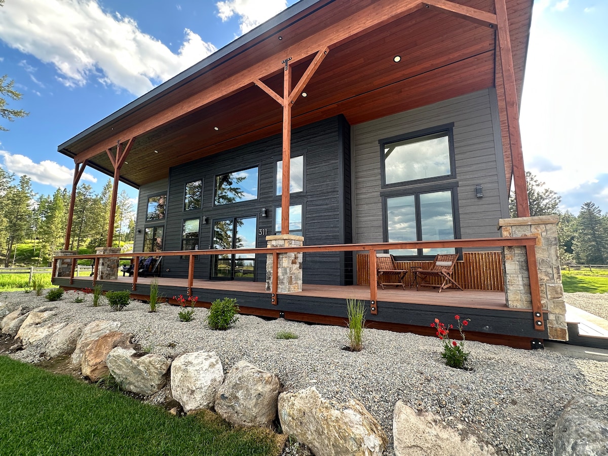 A modern home showcases a covered front deck with a wooden railing. Comfortable seating is positioned against large windows, inviting natural light. Stone accents and landscaping with gravel and small flowers facilitate a seamless connection with the surrounding nature.